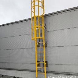 Tomasz Mech - Żółta drabina z koszem ochronnym zamontowana na szarej ścianie budynku. Widoczne elementy mocujące. Ujęcie od dołu, pochmurne niebo w tle.