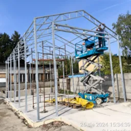 Montaż szkieletu metalowego hali: ekipa na podnośniku układa konstrukcję z profili, widoczne spawy, w tle budynek i zieleń. Prace w toku, słoneczny dzień.