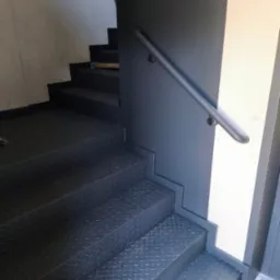 Metalowe schody z poręczą, pomalowane na ciemny kolor. Ryflowana powierzchnia stopni zapewnia antypoślizgowość. Widoczne mocowania poręczy do ściany.