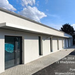 Tomasz Mech - Nowoczesny budynek z aluminiowymi zadaszeniami nad oknami i drzwiami. Szara stolarka okienna i drzwiowa. Ujęcie z zewnątrz, w słoneczny dzień.