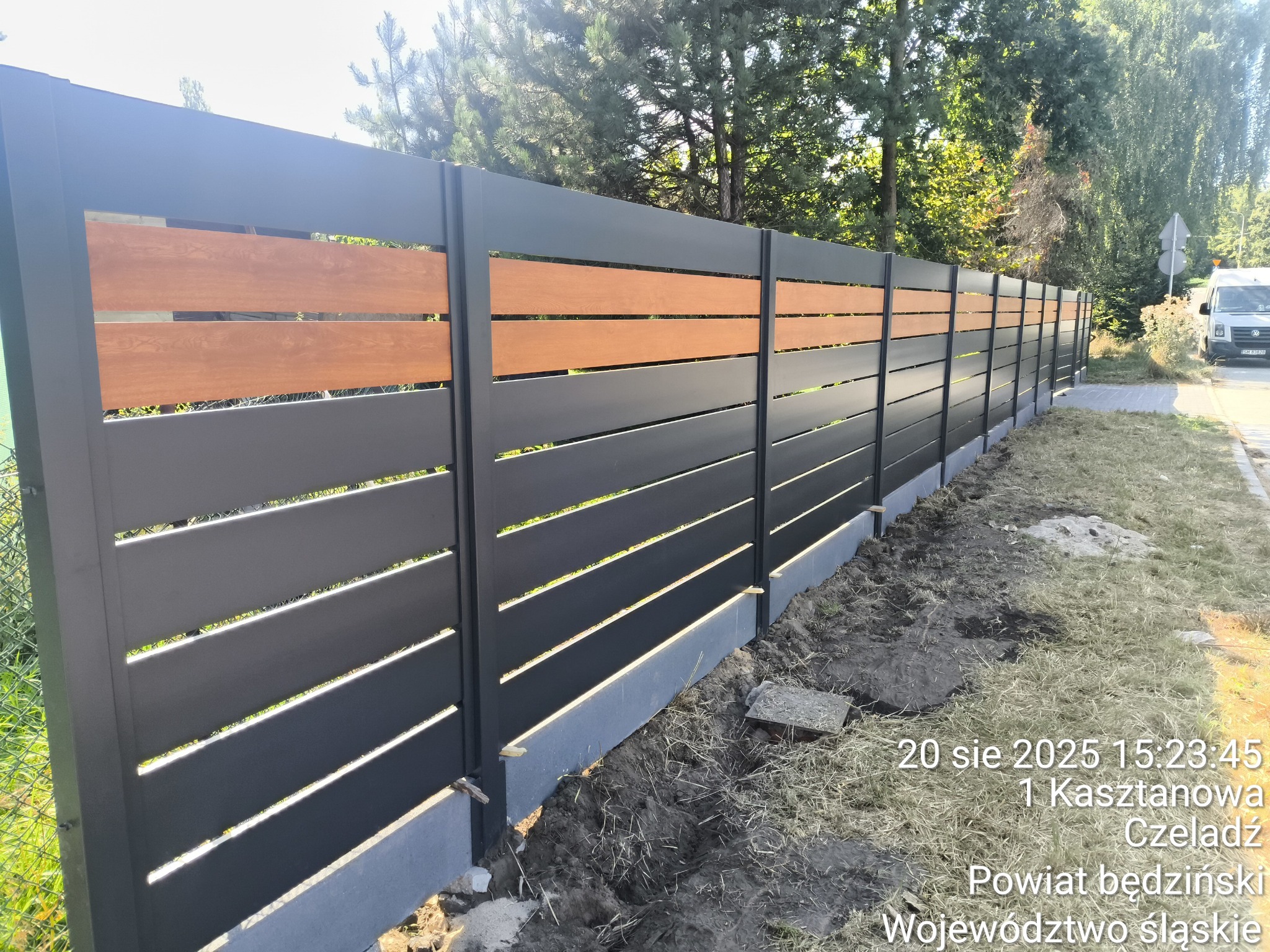 Nowoczesne ogrodzenie panelowe z poziomymi, grafitowymi przęsłami i drewnianymi akcentami. Widoczne słupki i podmurówka. Tło stanowi zieleń drzew i krzewów.