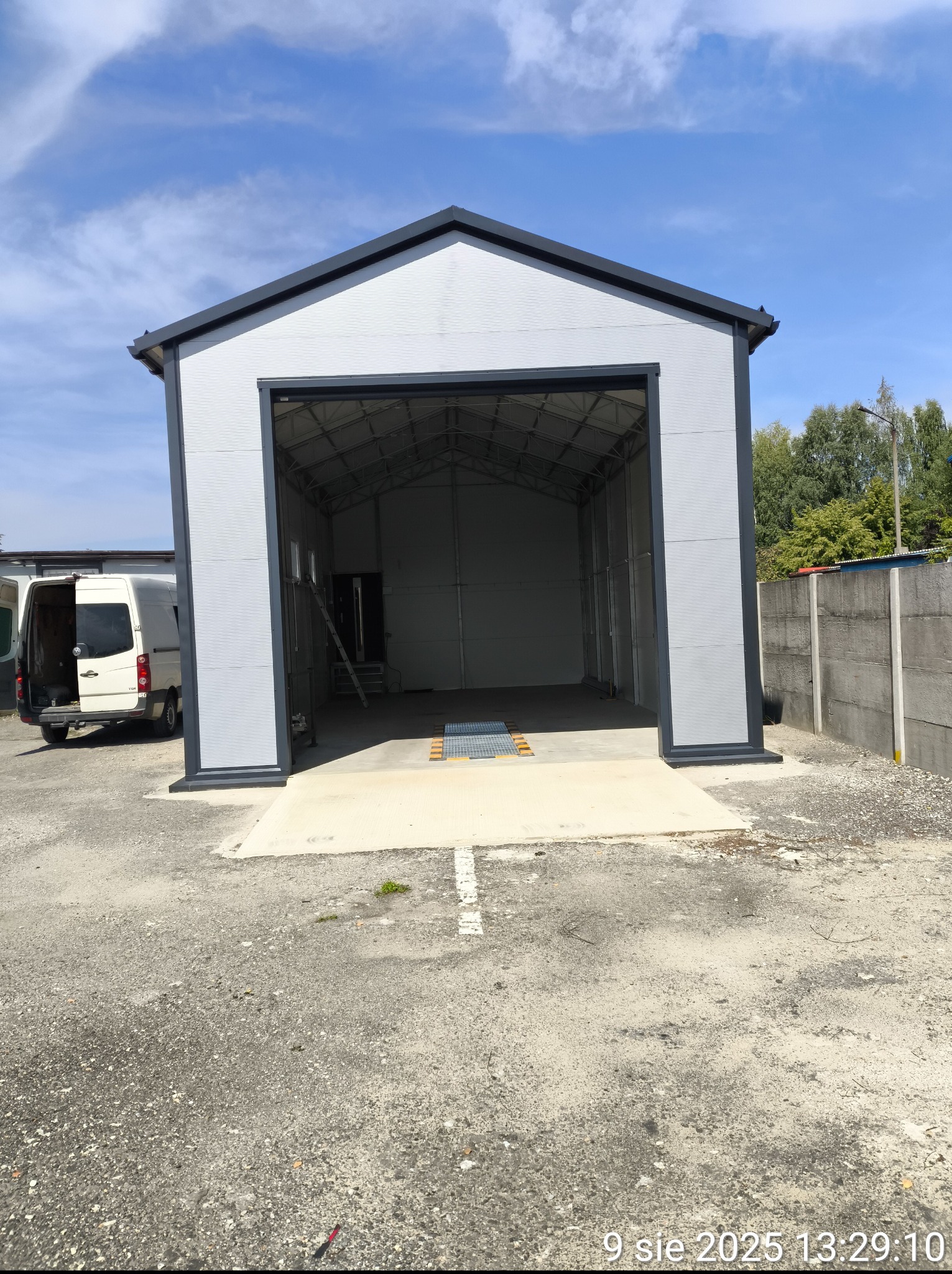 Szara, metalowa hala z otwartymi wrotami, widoczna drabina wewnątrz. Na zewnątrz zaparkowany biały bus, obok betonowe ogrodzenie. Błękitne niebo w tle.