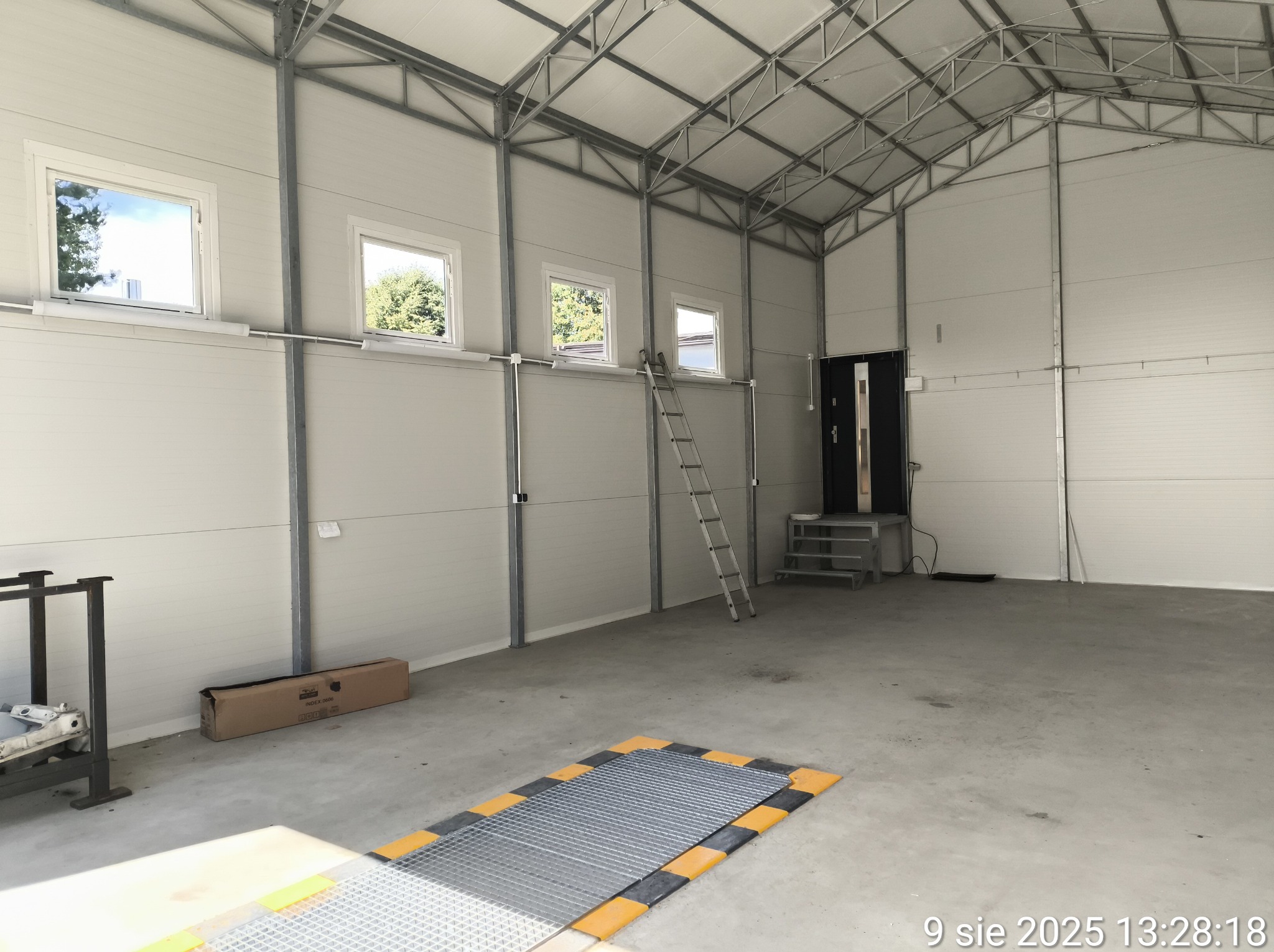 Wnętrze hali z metalową konstrukcją dachu i panelowymi ścianami. Widoczne okna, drabina oparta o ścianę, kratka pomostowa w podłodze i metalowy wózek.