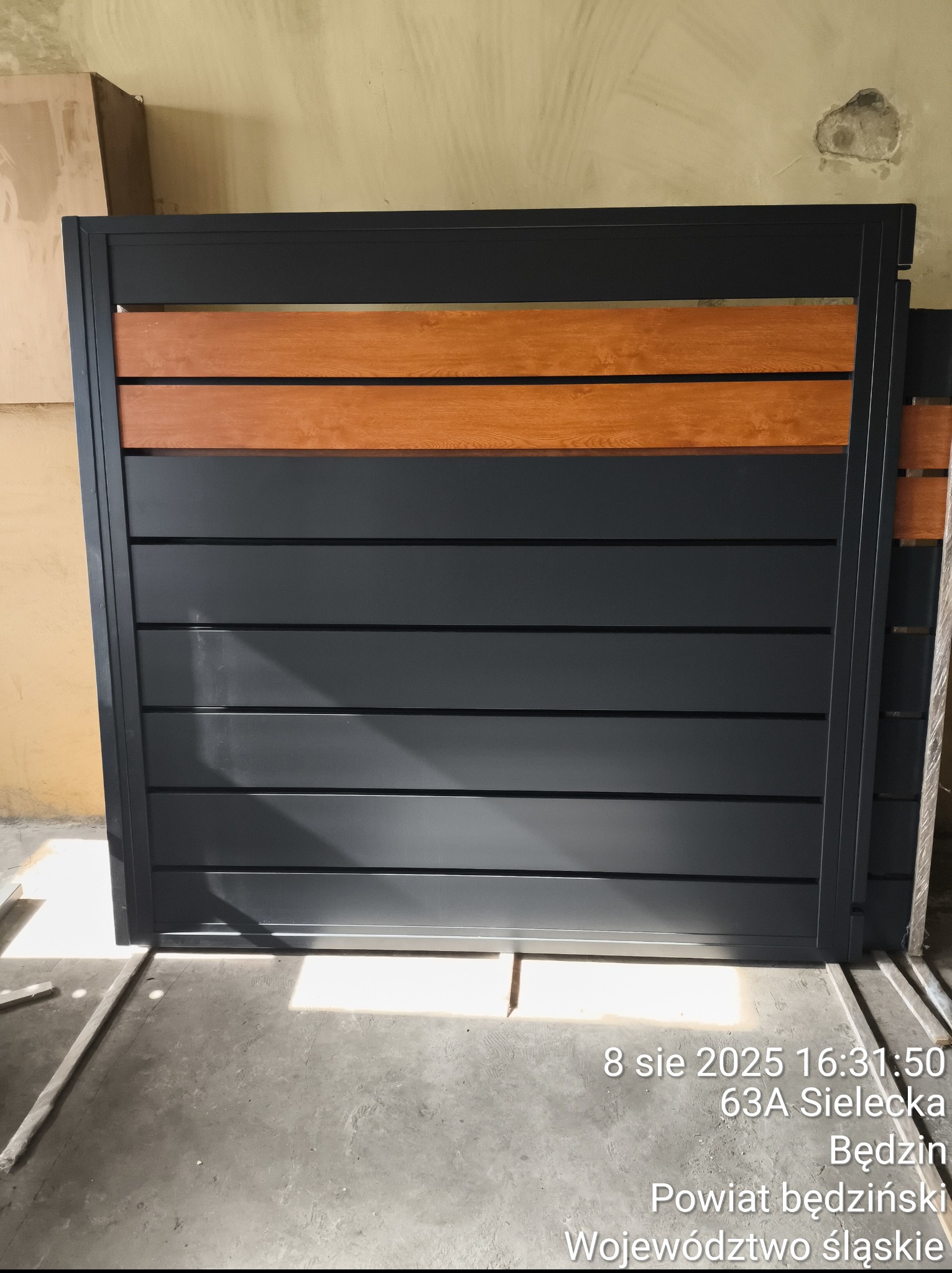 Nowoczesna, przesuwna furtka z szarych, metalowych paneli i drewnianych akcentów. Widoczne detale konstrukcyjne i elementy mocujące. Furtka w trakcie montażu.