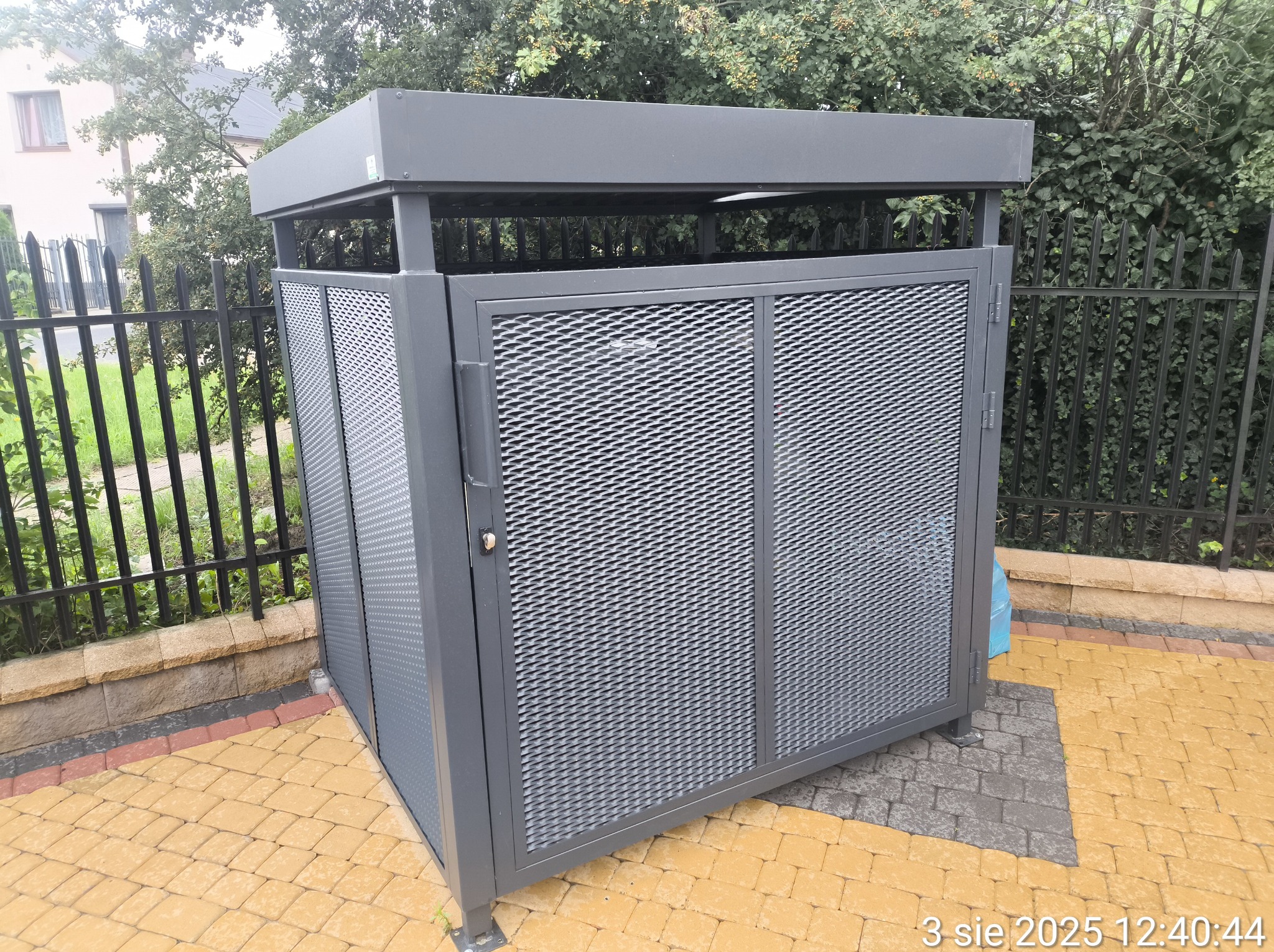 Szara, metalowa obudowa na śmietniki z ażurowymi drzwiami, na kostce brukowej. W tle czarne ogrodzenie i zieleń.