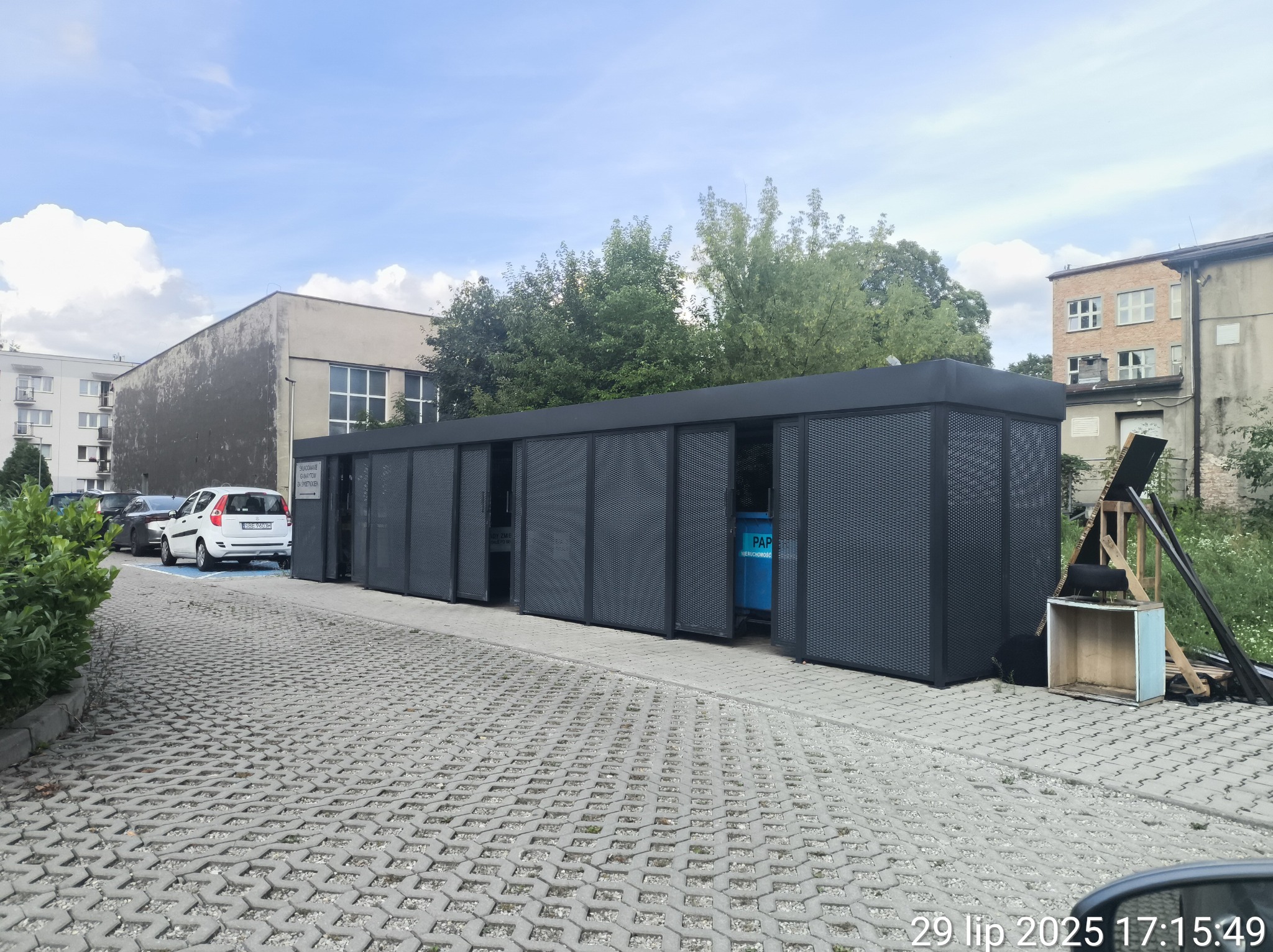 Metalowa konstrukcja osłaniająca pojemniki na odpady na zewnątrz, z ażurowymi ścianami i przesuwanymi drzwiami, na tle budynków i zieleni.