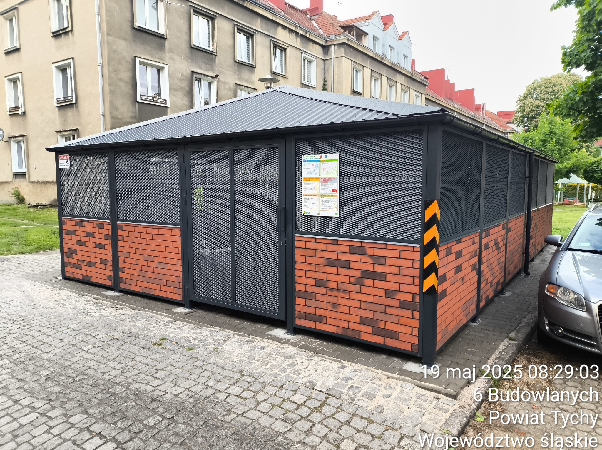 Zadaszona konstrukcja z metalową siatką i ceglaną podstawą, prawdopodobnie wiata śmietnikowa, z żółto-czarnymi pasami ostrzegawczymi na słupie, na tle budynków mieszkalnych.