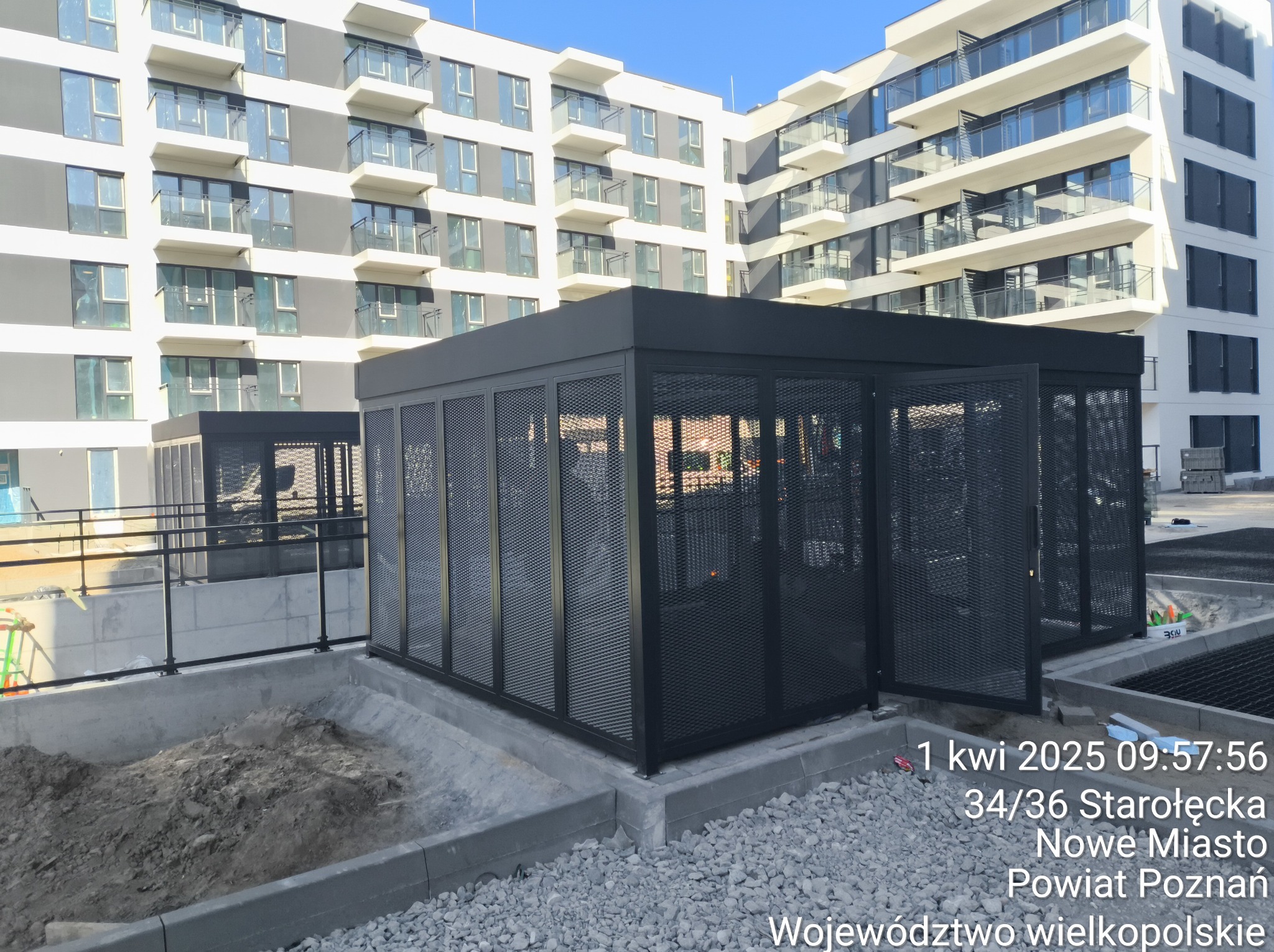 Nowoczesna, ażurowa konstrukcja metalowa w kolorze czarnym, prawdopodobnie na balkonie lub tarasie, z budynkiem w tle. Otwarta brama z siatki.