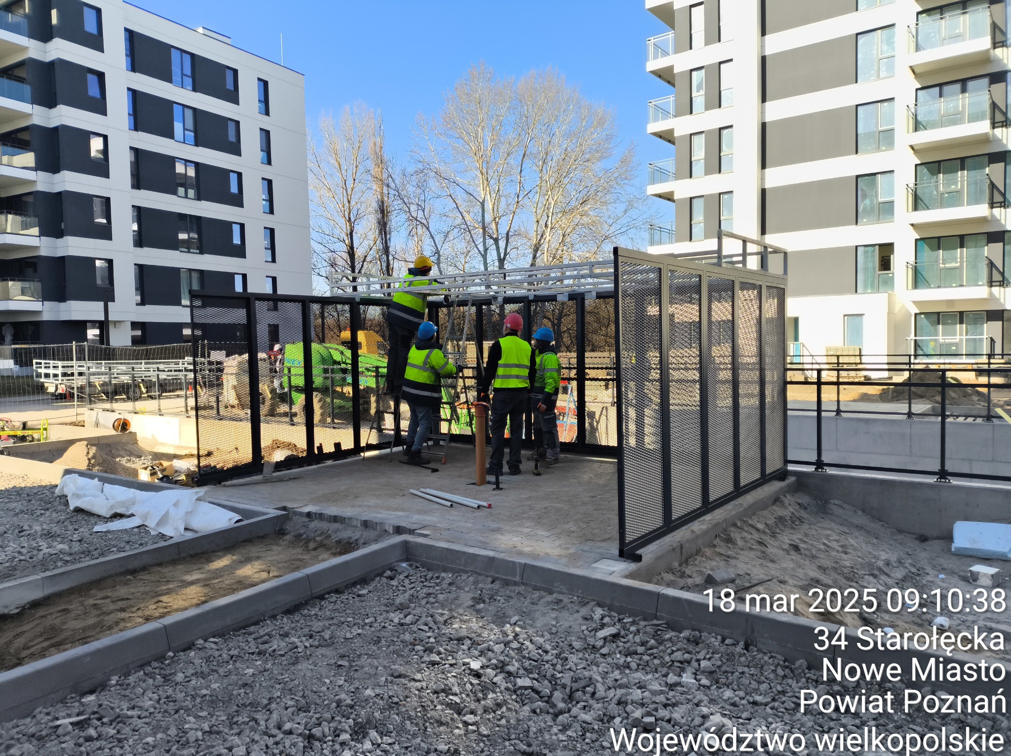 Montaż ażurowej konstrukcji przy nowym budynku. Pracownicy w odblaskowych kamizelkach. Widoczne elementy konstrukcyjne i narzędzia. Słoneczny dzień.