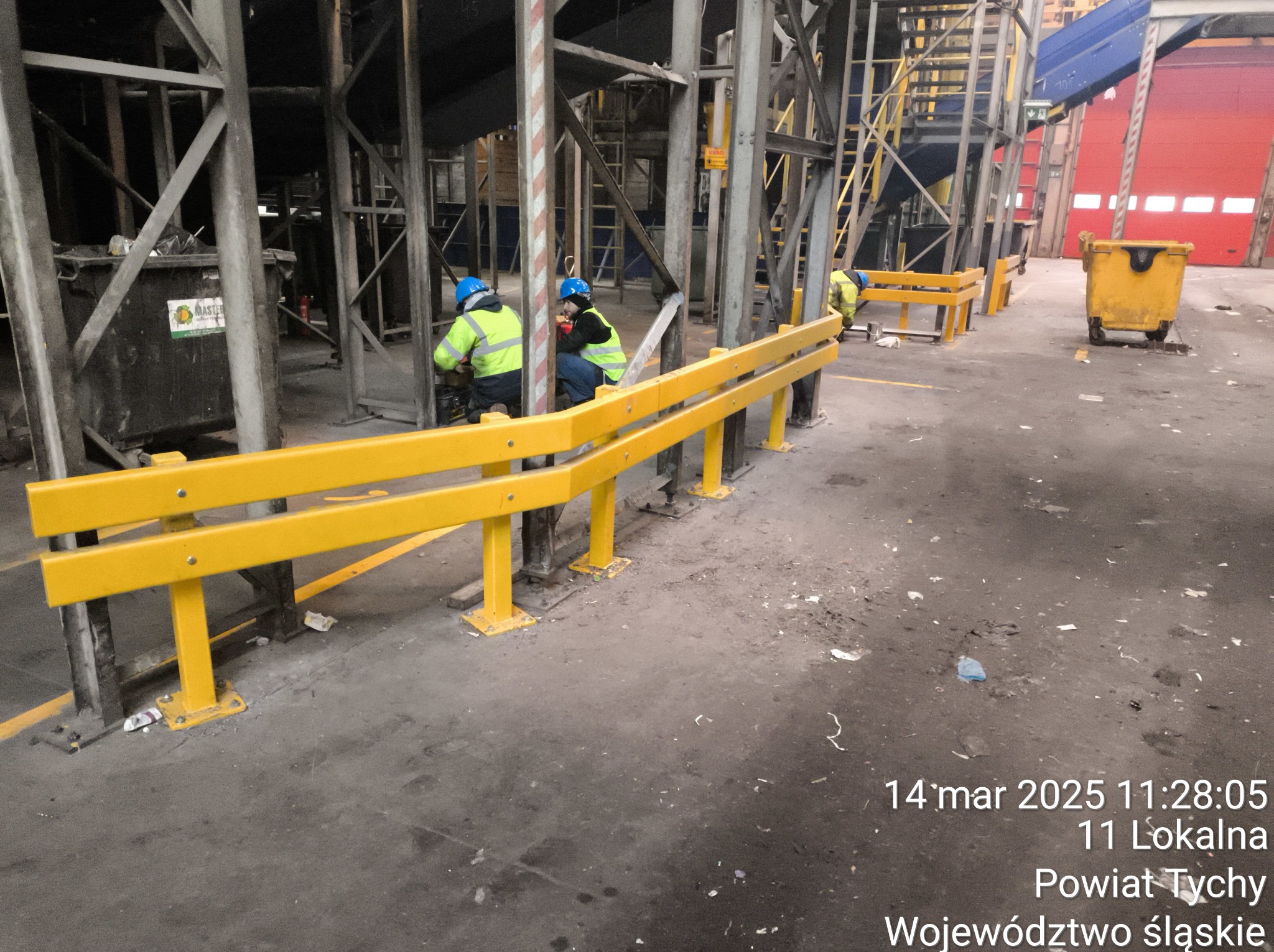 Żółta barierka ochronna z metalu w hali przemysłowej, pracownicy w kaskach wykonują prace montażowe. Widoczne elementy konstrukcyjne i wózek na śmieci.