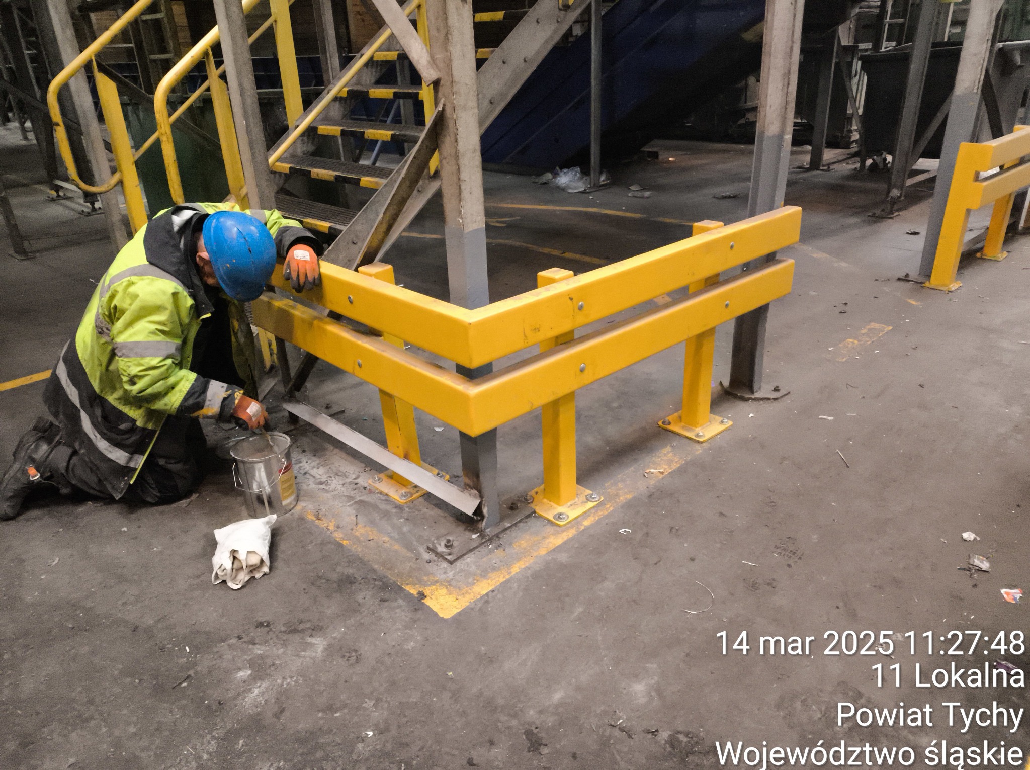 Pracownik w kasku maluje żółtą farbą metalową konstrukcję ochronną w hali przemysłowej. Widoczne schody, podłoga z zabrudzeniami i datownik.