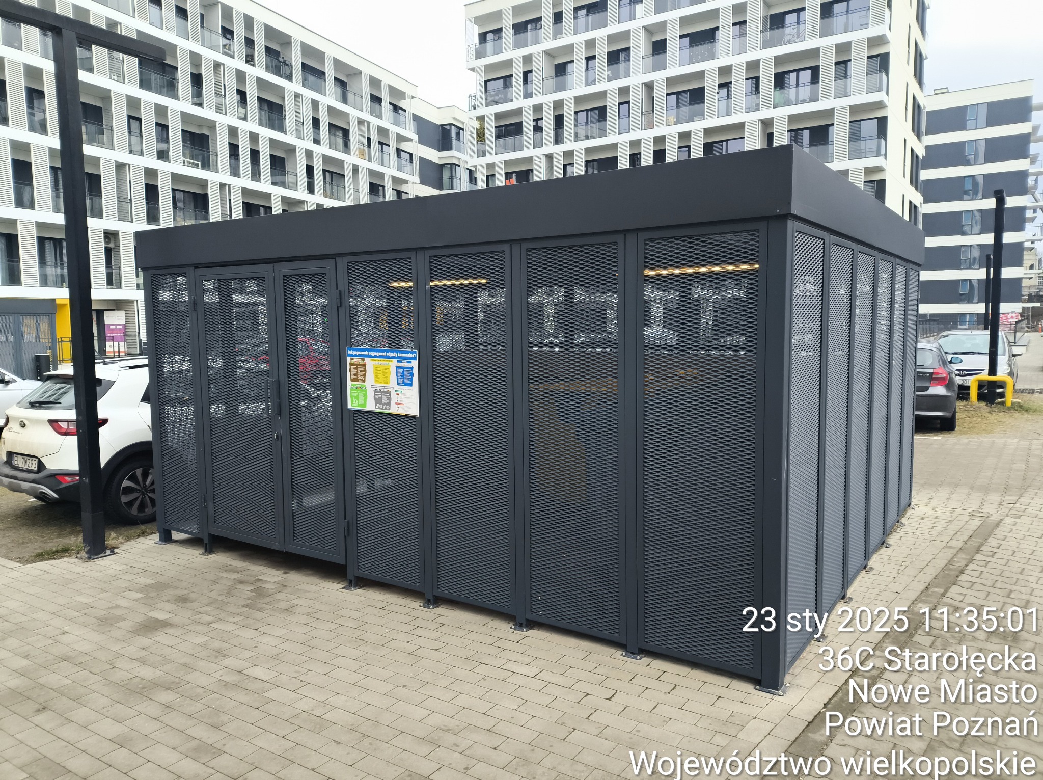 Metalowa konstrukcja z perforowanymi ścianami, prawdopodobnie wiata rowerowa, na tle nowoczesnego budynku mieszkalnego. Widoczny fragment chodnika i zaparkowane samochody.