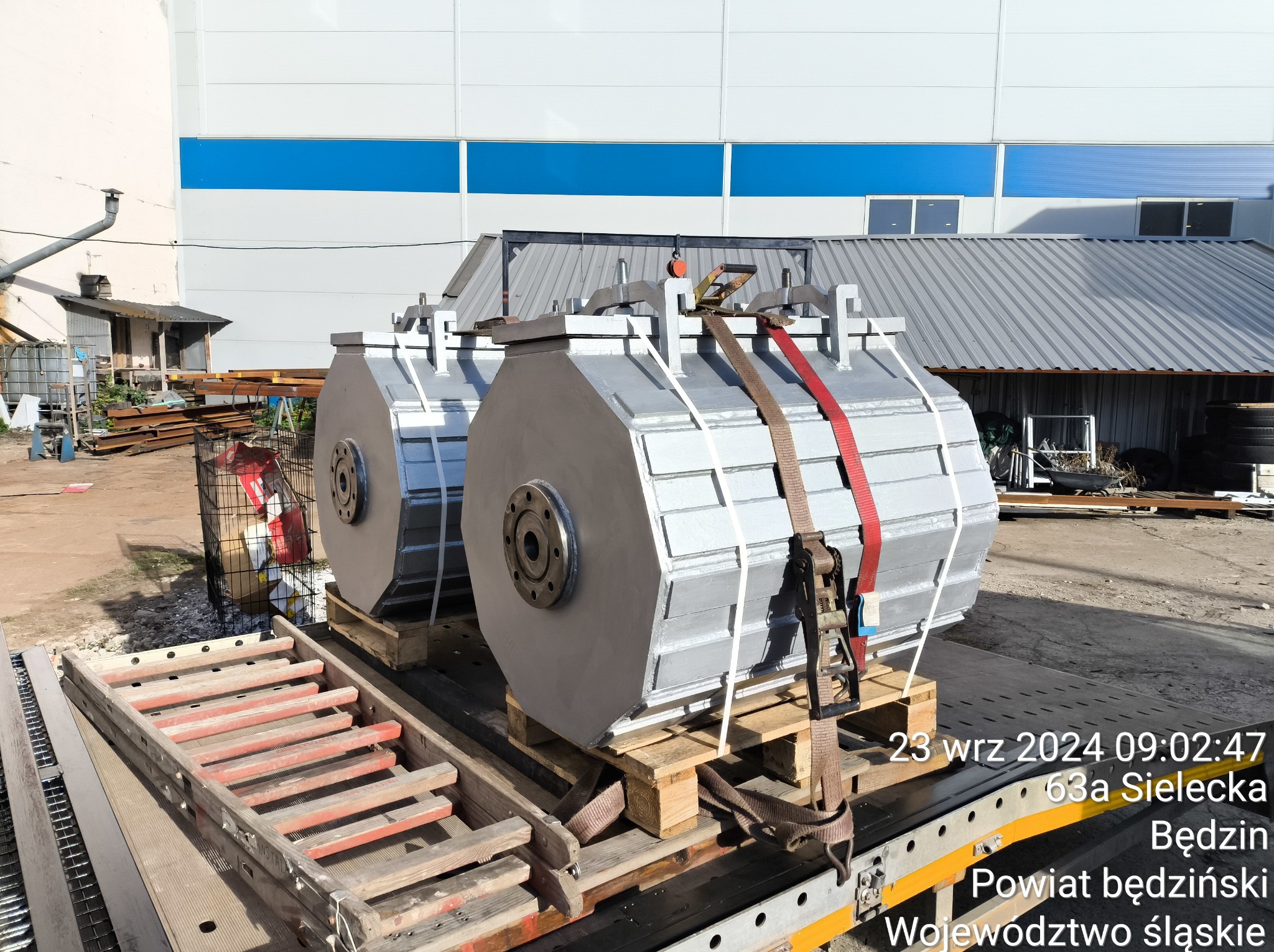 Dwa duże, srebrne elementy metalowe o cylindrycznym kształcie, zabezpieczone pasami transportowymi, na platformie pojazdu. Widoczne detale konstrukcyjne i elementy złączne.