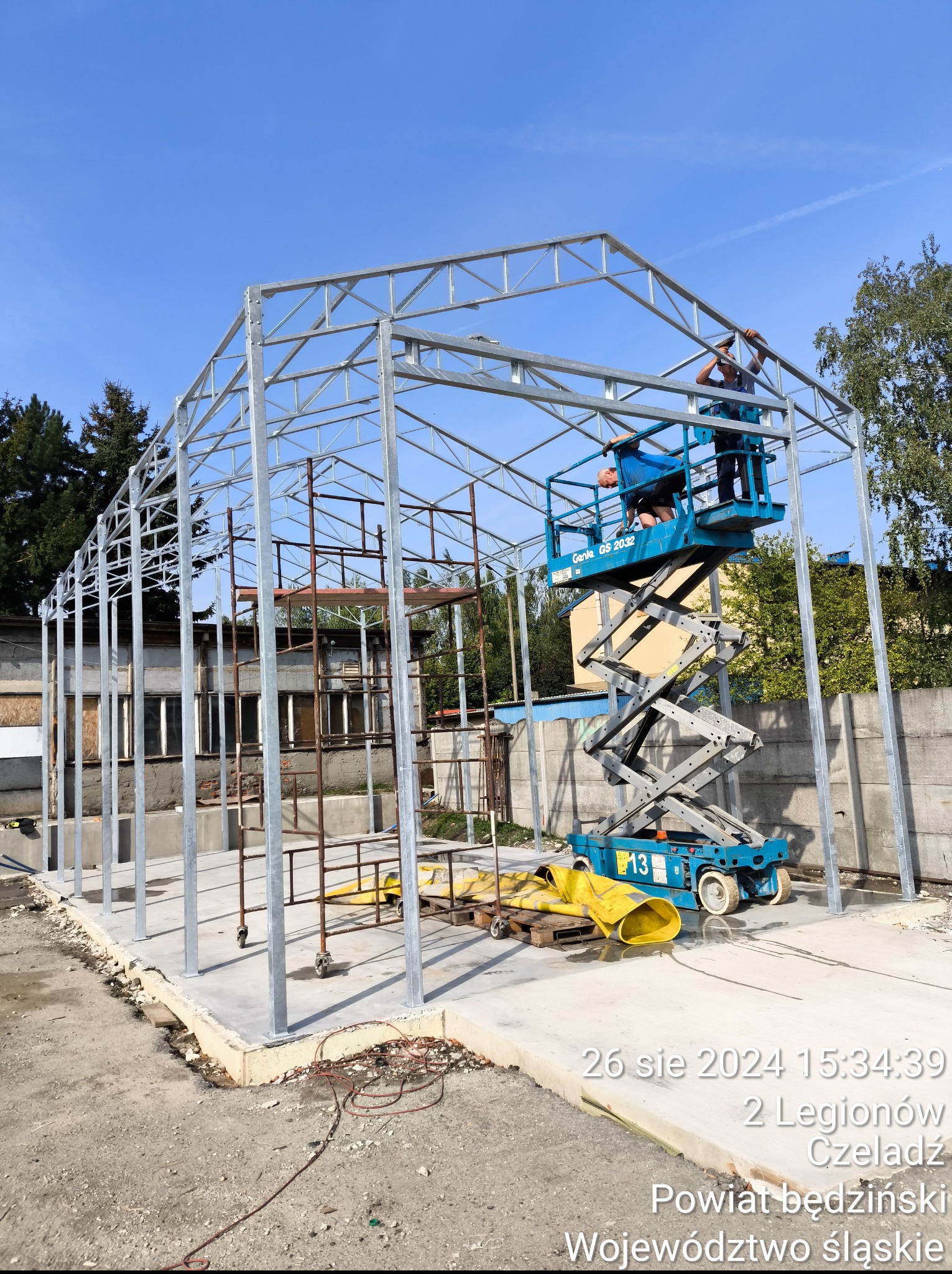 Montaż szkieletu metalowego hali: ekipa na podnośniku układa konstrukcję z profili, widoczne spawy, w tle budynek i zieleń. Prace w toku, słoneczny dzień.