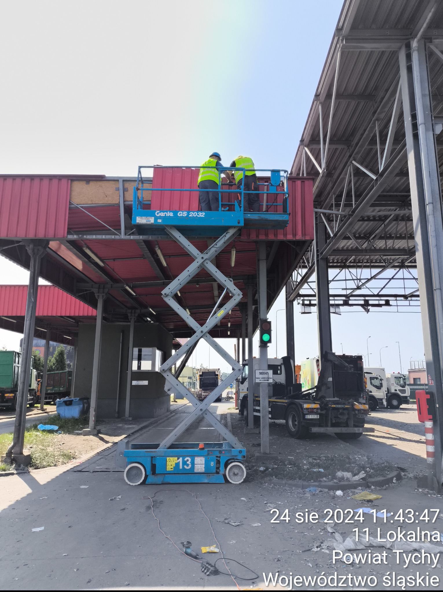 Pracownicy na podnośniku montują elementy konstrukcyjne na zadaszeniu, prawdopodobnie przy punkcie kontrolnym. Widoczne ciężarówki i sygnalizacja świetlna.