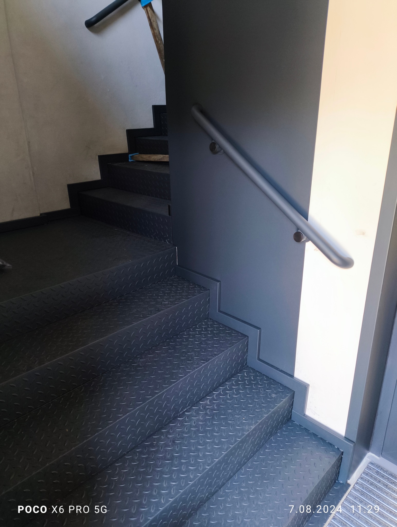 Metalowe schody z poręczą, pomalowane na ciemny kolor. Ryflowana powierzchnia stopni zapewnia antypoślizgowość. Widoczne mocowania poręczy do ściany.