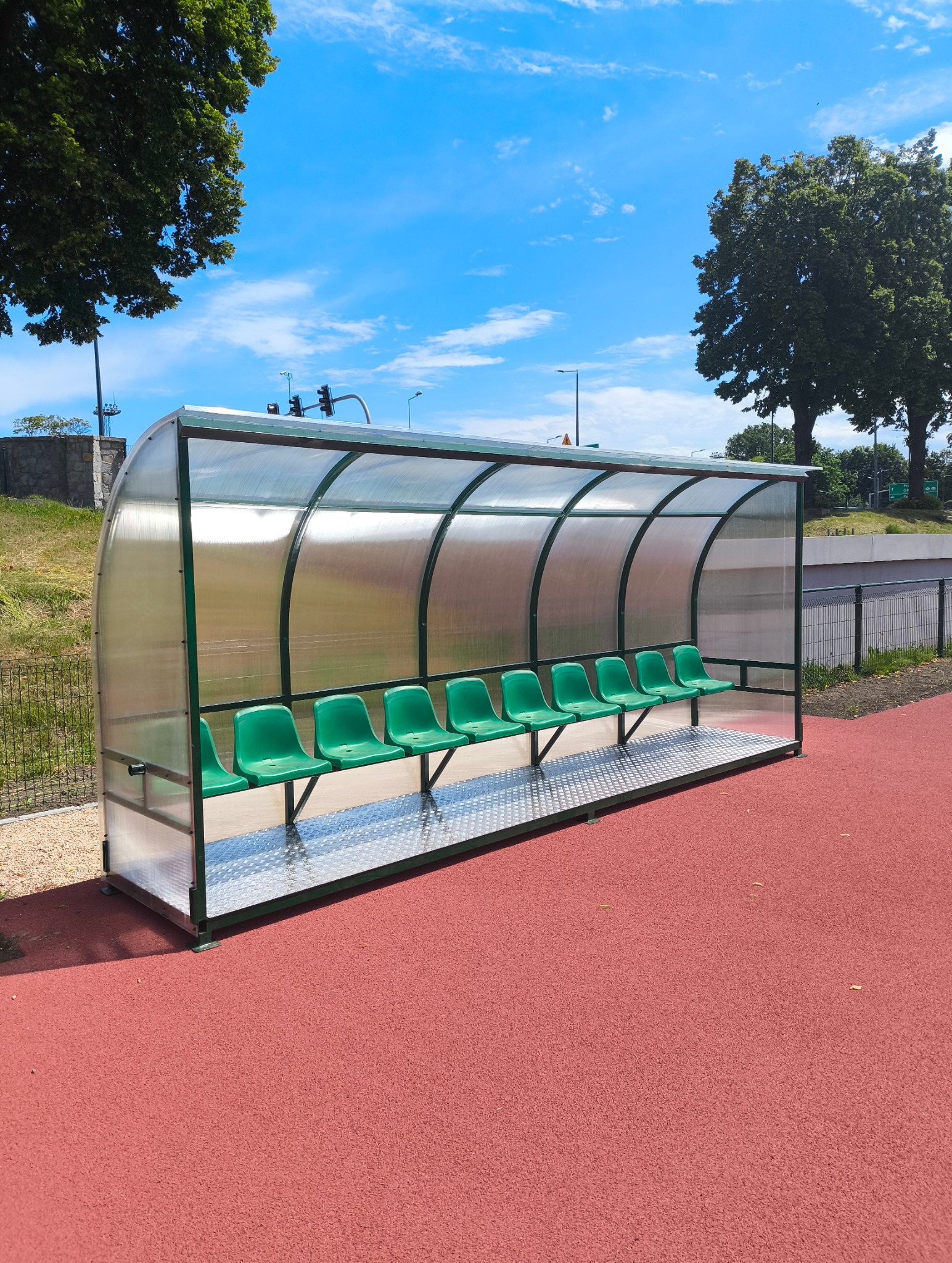 Zadaszona wiata z zielonymi siedzeniami na czerwonej nawierzchni boiska, otoczona zielenią i błękitnym niebem. Konstrukcja metalowa z przezroczystym zadaszeniem.