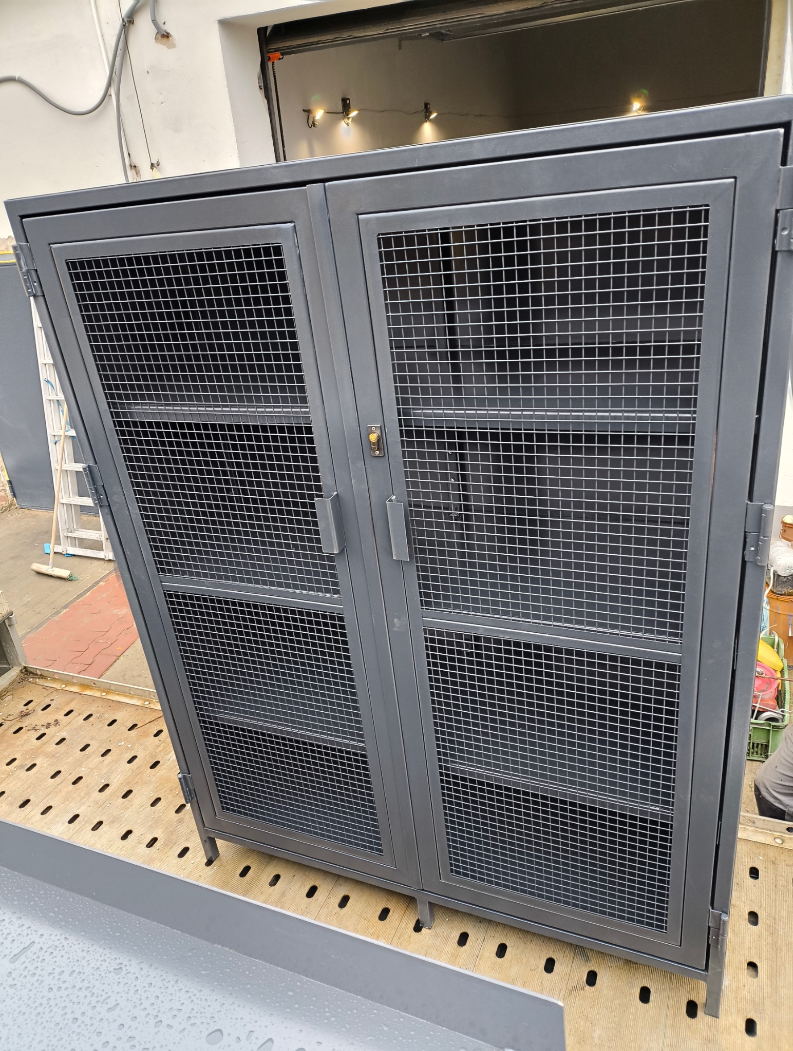 Metalowa szafka z siatki, dwudrzwiowa, zamykana na zamek, na tle otwartej bramy garażowej i drabiny. Szary kolor, prosta konstrukcja.