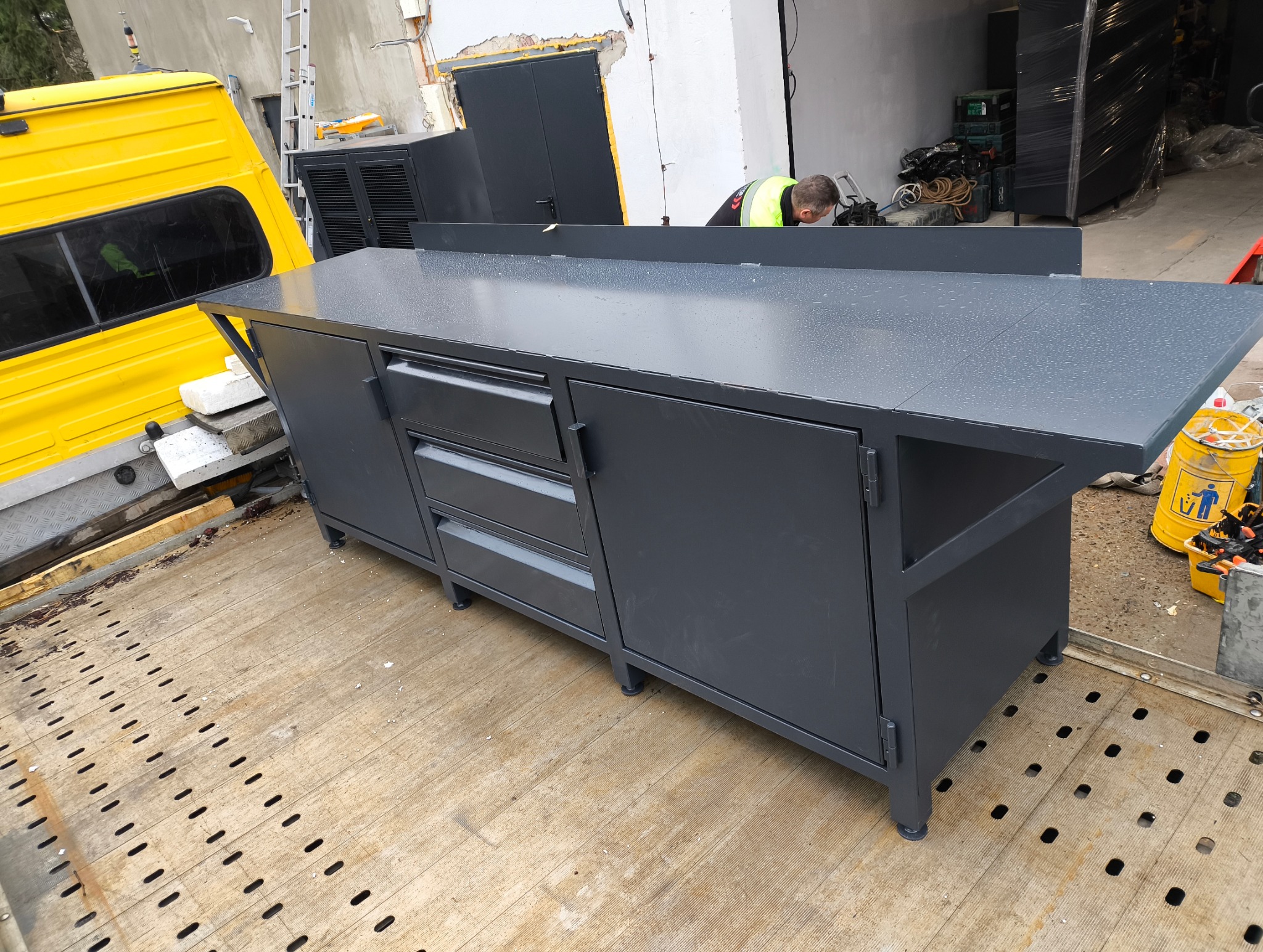 Szara, metalowa szafka warsztatowa z blatem, trzema szufladami i szafkami, stojąca na platformie transportowej. Widoczny fragment żółtego samochodu dostawczego.