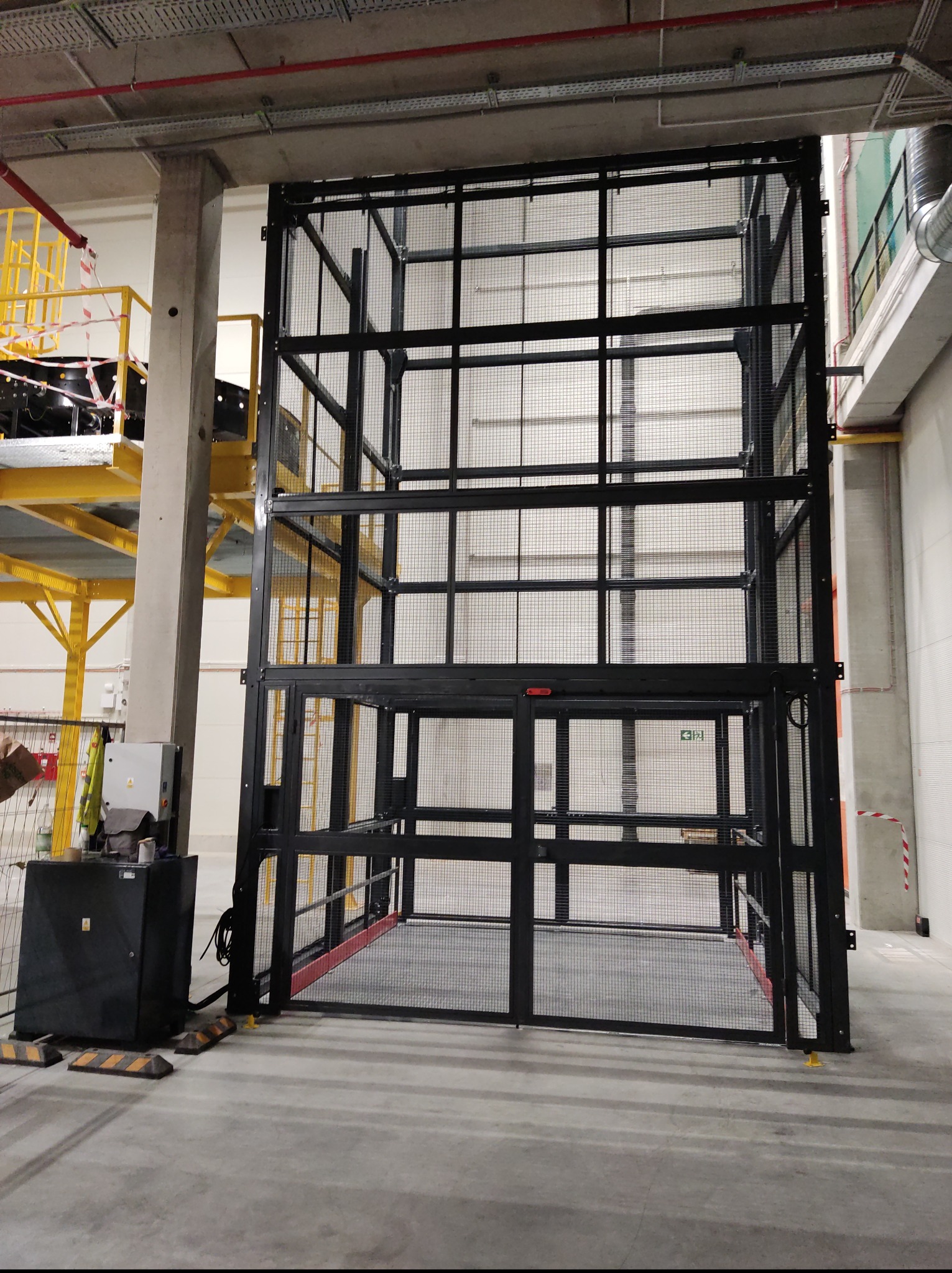 Wytrzymała, czarna konstrukcja z siatki, przypominająca klatkę towarową, w industrialnym wnętrzu. Widoczne elementy mocujące i podesty, surowy beton na podłodze i ścianach.