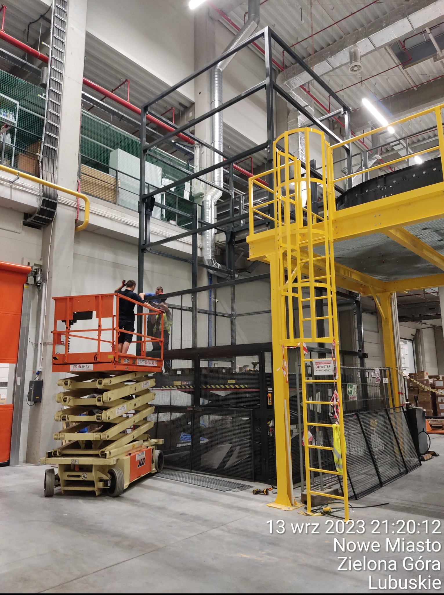 Montaż konstrukcji stalowej w hali magazynowej z użyciem podnośnika nożycowego. Widoczne elementy instalacji i oświetlenia. Prace na wysokościach w trakcie realizacji.