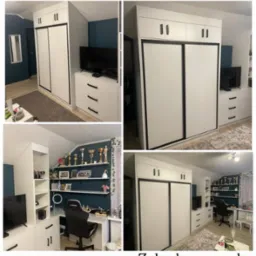 Biała szafa wnękowa z przesuwnymi drzwiami i szafkami górnymi, wkomponowana w pokój z biurkiem i półkami. Widoczne trofea i dekoracje, ciemna ściana w tle.