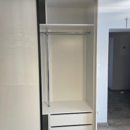 Mateusz Ratajczak - Wnętrze szafy wnękowej z przesuwnymi drzwiami i szufladami, biała z czarnymi obramowaniami, widoczny drążek na ubrania. W tle fragment pokoju.