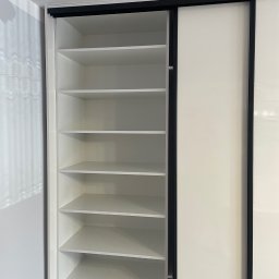 Mateusz Ratajczak - Biała szafa wnękowa z przesuwnymi drzwiami i czarnymi obramowaniami, widok na puste półki. Minimalistyczny design, jasne wnętrze szafy.