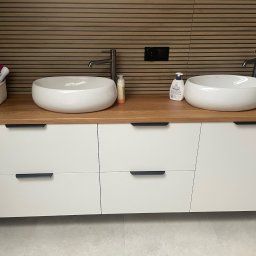 Mateusz Ratajczak - Nowoczesna szafka łazienkowa z dwoma okrągłymi umywalkami nablatowymi, drewnianym blatem i białymi frontami szuflad na tle brązowej ściany z poziomych paneli.