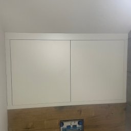 Mateusz Ratajczak - Białe szafki nad zabudową WC z drewnopodobnymi płytkami, widoczne przyłącza wodne. Minimalistyczne rozwiązanie w łazience.