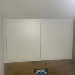 Białe szafki nad zabudową WC z drewnopodobnymi płytkami, widoczne przyłącza wodne. Minimalistyczne rozwiązanie w łazience.