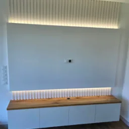 Biała zabudowa RTV z drewnianym blatem i oświetleniem LED, pionowe lamele dekoracyjne nad panelem ściennym. Nowoczesny design, widok na gniazdka.