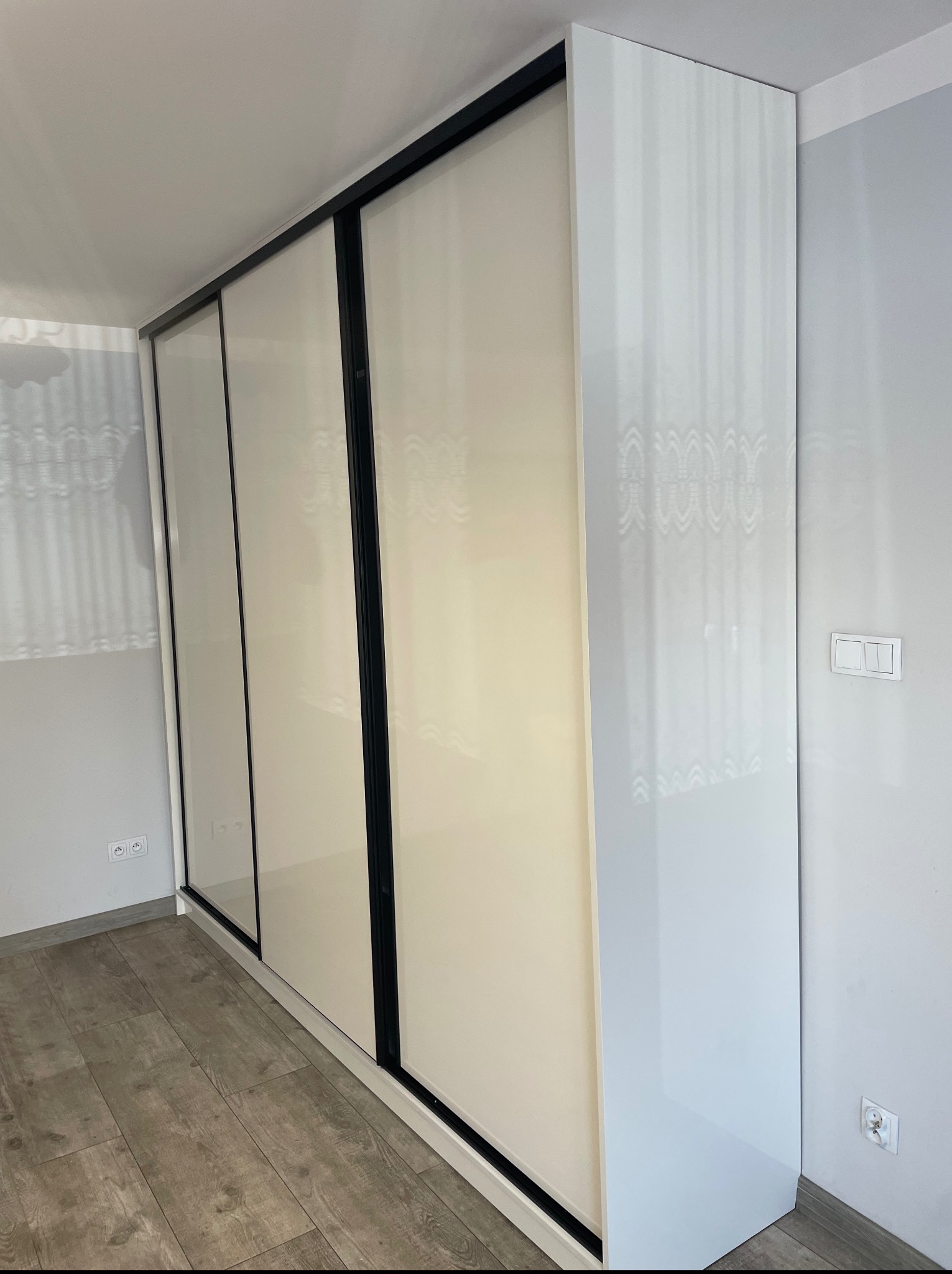 Biała szafa wnękowa z przesuwnymi drzwiami i czarnymi ramami, dopasowana do sufitu, z widocznymi gniazdkami elektrycznymi i drewnianą podłogą.