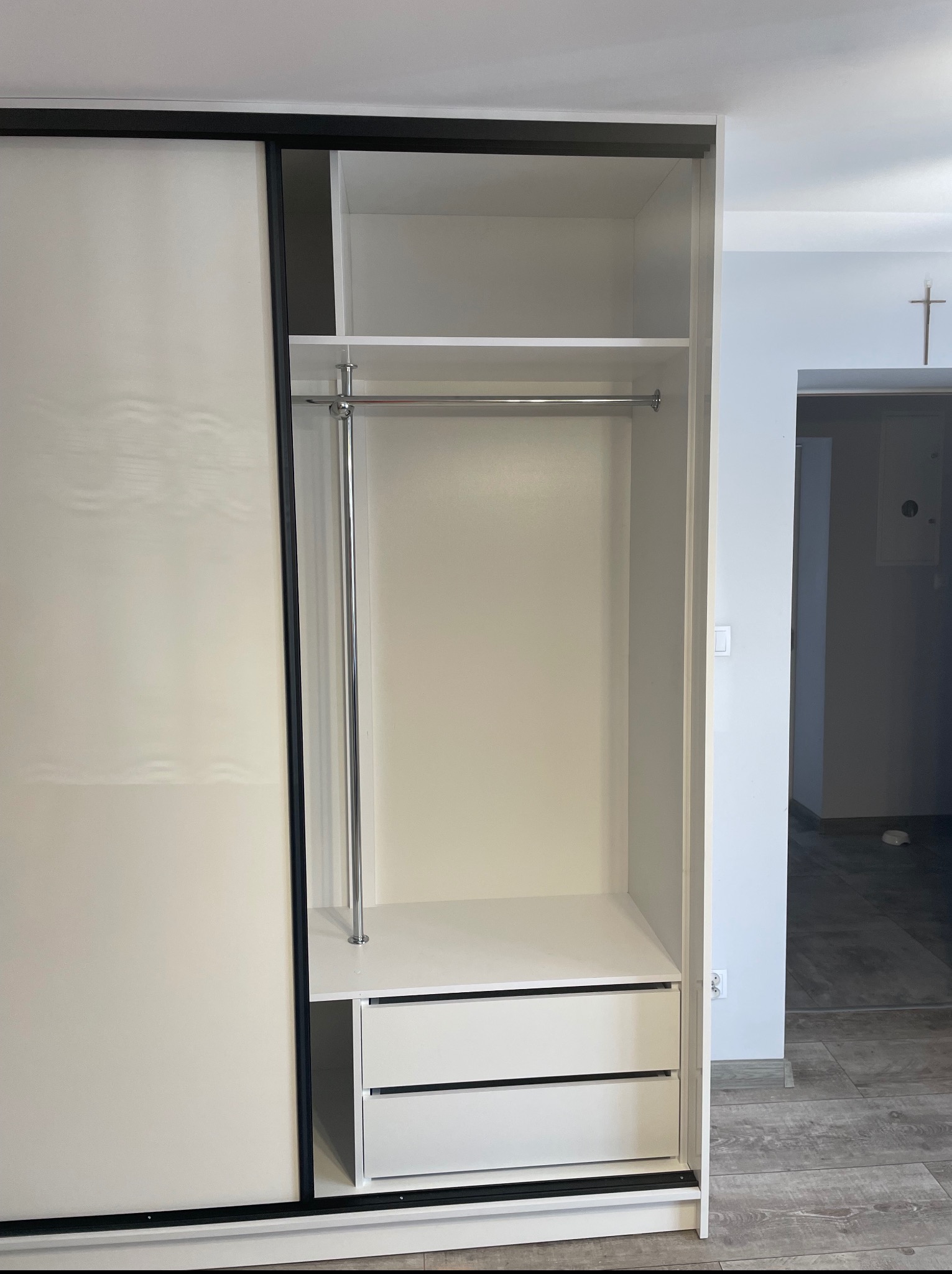 Wnętrze szafy wnękowej z przesuwnymi drzwiami i szufladami, biała z czarnymi obramowaniami, widoczny drążek na ubrania. W tle fragment pokoju.