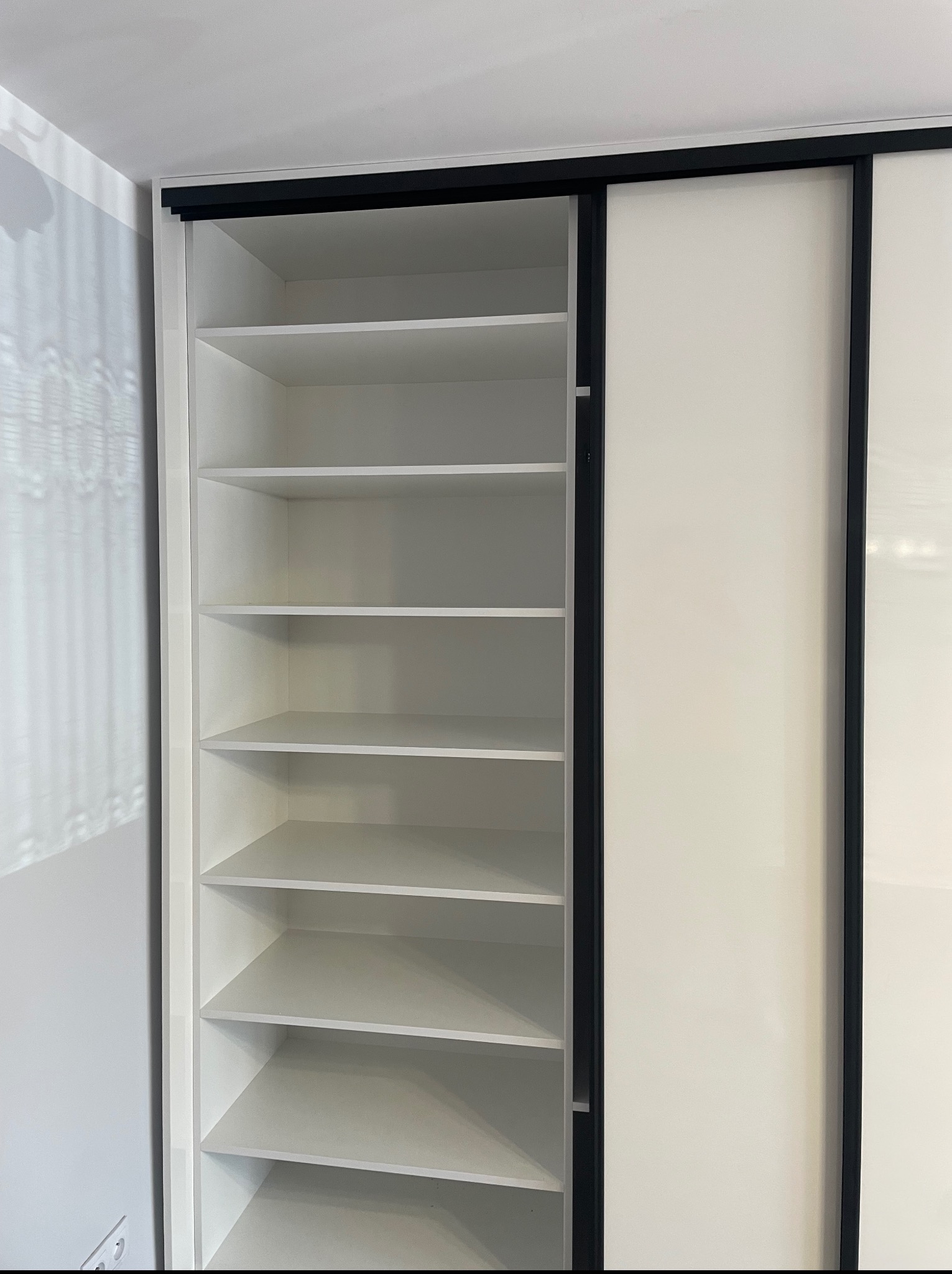 Biała szafa wnękowa z przesuwnymi drzwiami i czarnymi obramowaniami, widok na puste półki. Minimalistyczny design, jasne wnętrze szafy.