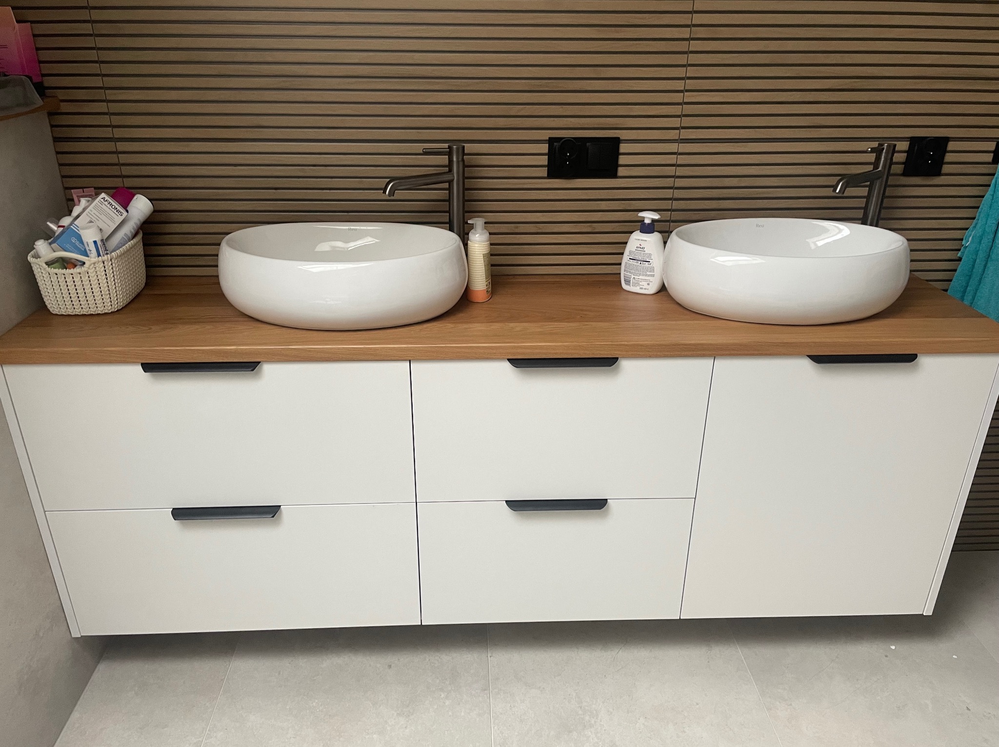 Nowoczesna szafka łazienkowa z dwoma okrągłymi umywalkami nablatowymi, drewnianym blatem i białymi frontami szuflad na tle brązowej ściany z poziomych paneli.