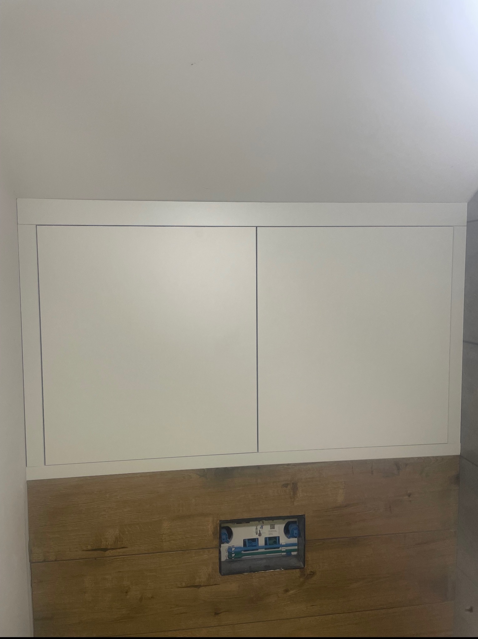 Białe szafki nad zabudową WC z drewnopodobnymi płytkami, widoczne przyłącza wodne. Minimalistyczne rozwiązanie w łazience.