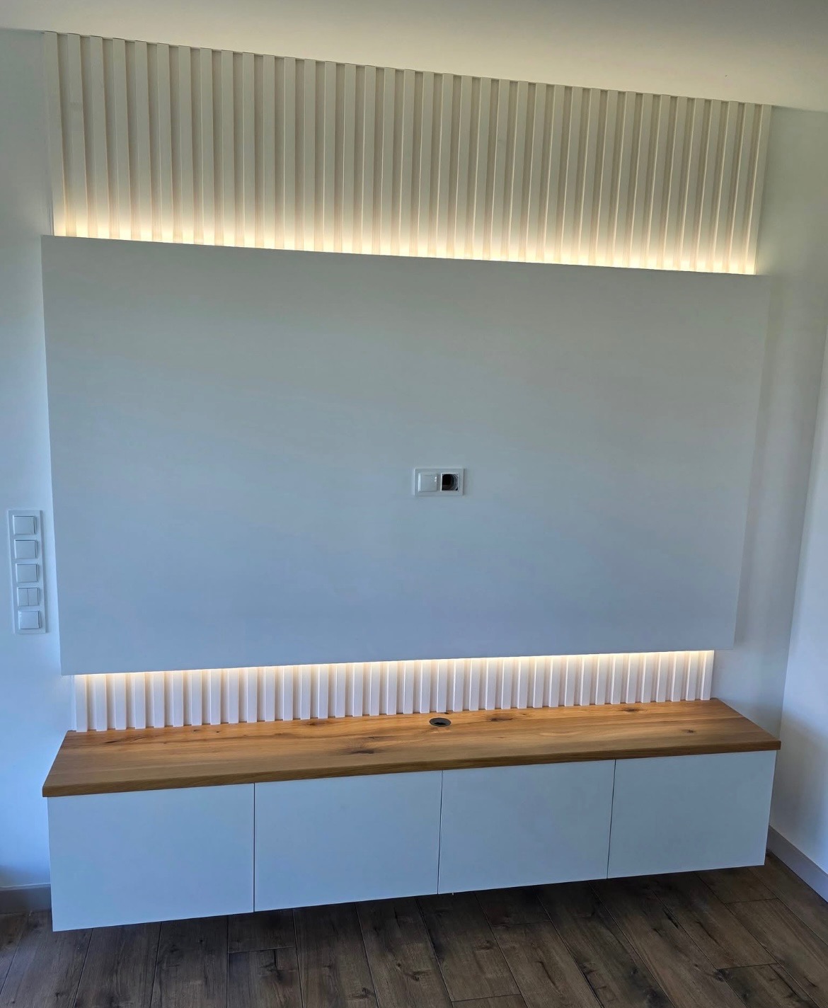 Biała zabudowa RTV z drewnianym blatem i oświetleniem LED, pionowe lamele dekoracyjne nad panelem ściennym. Nowoczesny design, widok na gniazdka.