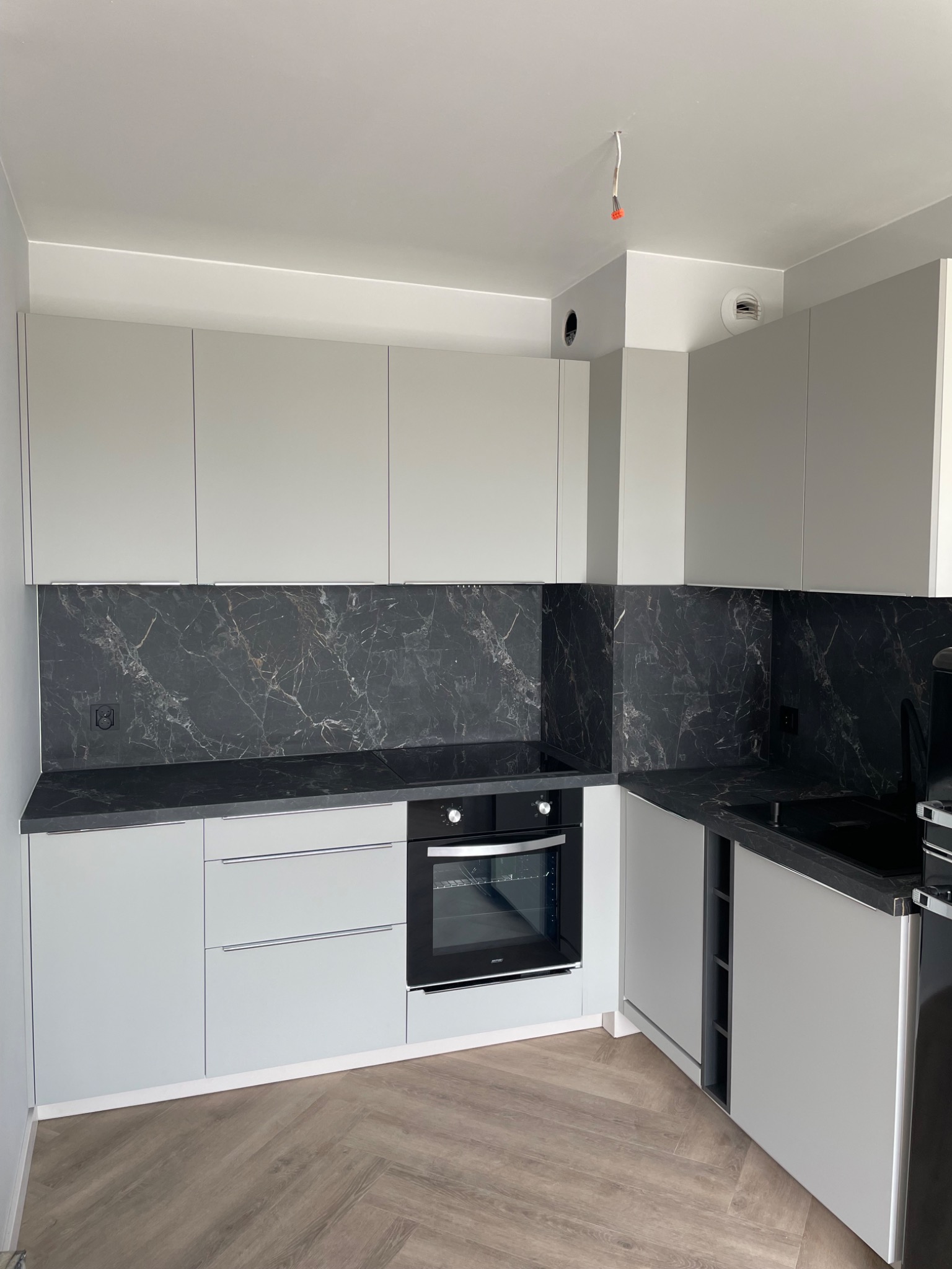Jasnoszara kuchnia z czarnym blatem i marmurowym panelem. Widoczny piekarnik i drewniana podłoga w jodełkę. Minimalistyczny design z oświetleniem punktowym.
