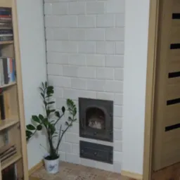 Biały piec kaflowy z żeliwnymi drzwiczkami, obok drewniana szafka z książkami i roślina doniczkowa, parkiet na podłodze, widok na drzwi.