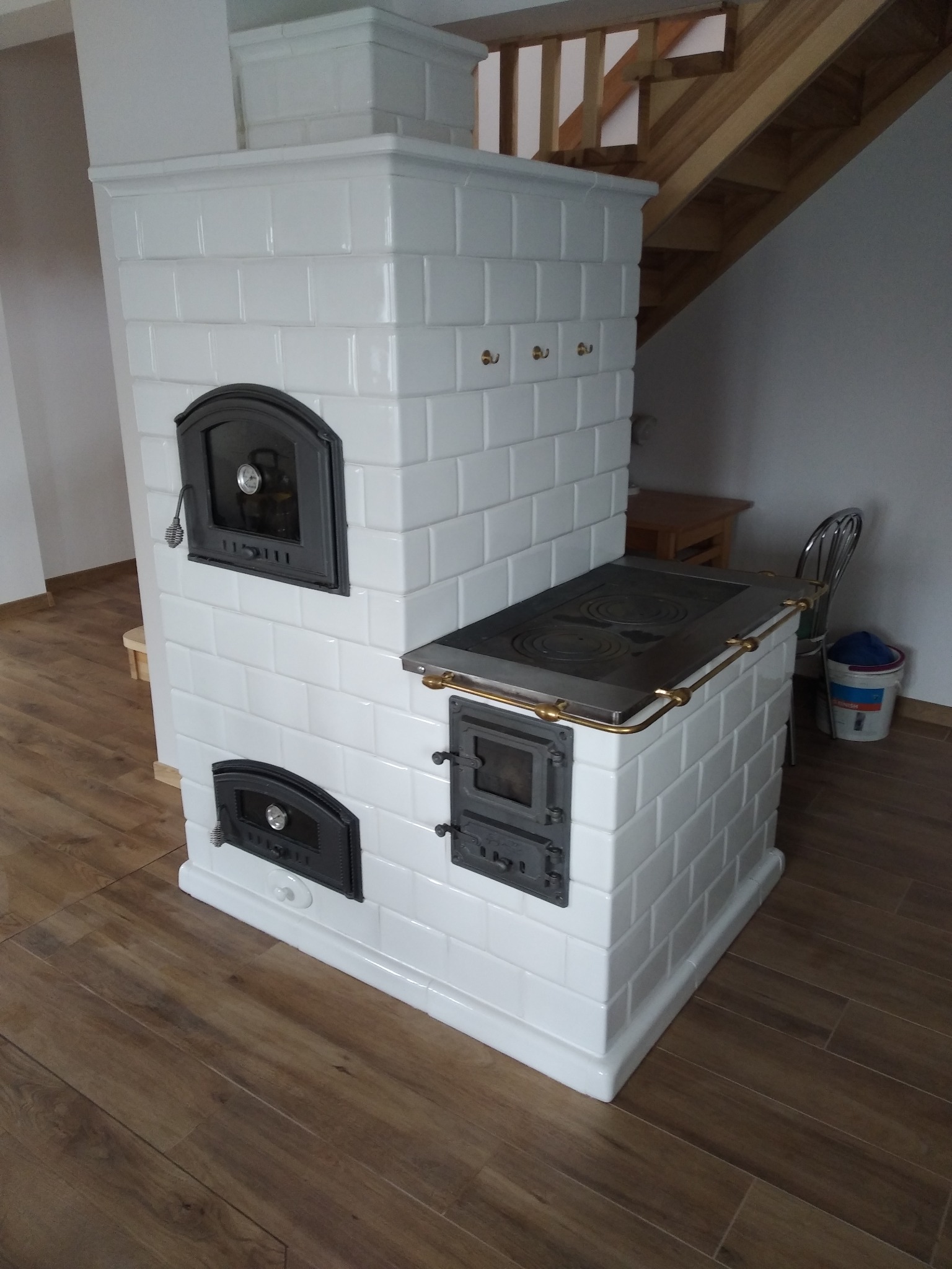 Biały piec kaflowy z czarnymi drzwiczkami na drewnianej podłodze. Widoczne fragmenty schodów i krzesła. Piec w stylu retro, z płytą grzewczą na górze.