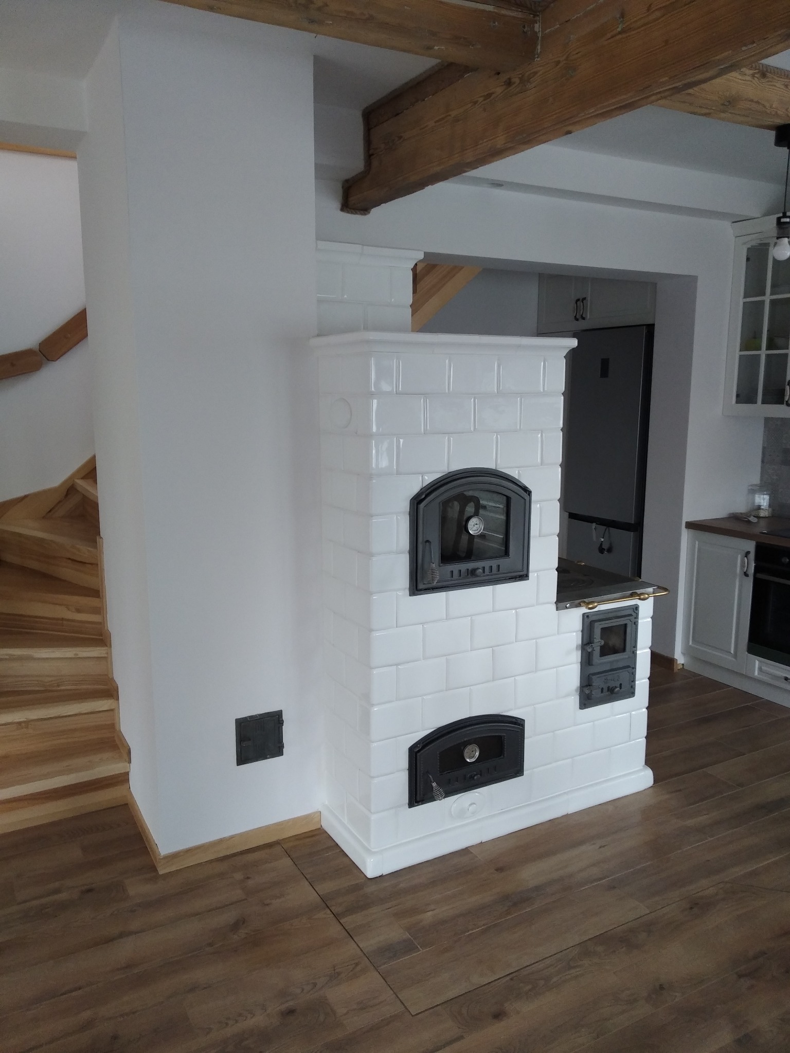 Biały piec kaflowy z czarnymi drzwiczkami, wkomponowany w nowoczesne wnętrze z drewnianymi schodami i belkami stropowymi. Widoczne detale wykończenia.