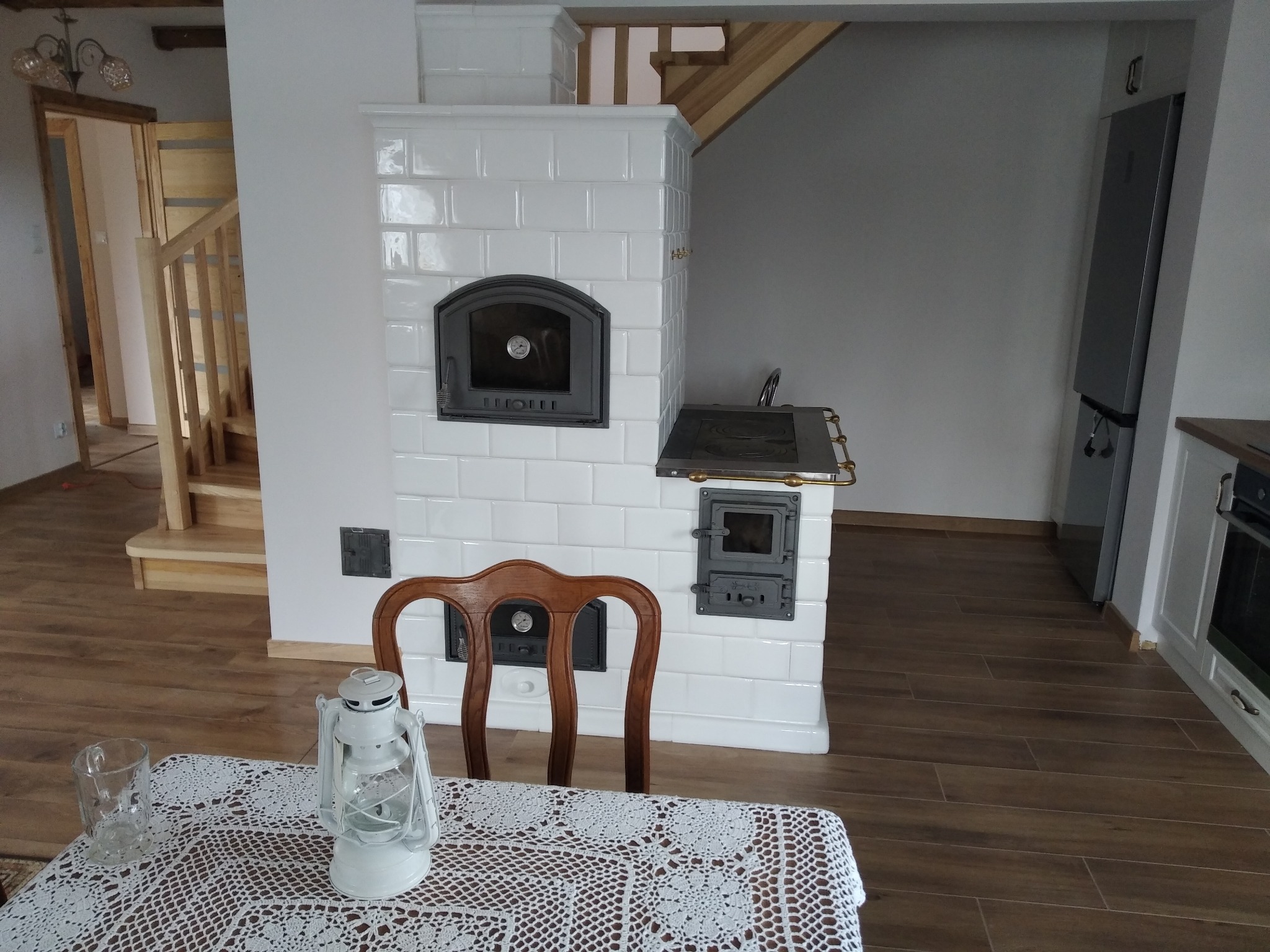 Biały piec kaflowy z żeliwnymi drzwiczkami w jasnym wnętrzu. Na pierwszym planie stół z koronkowym obrusem i lampą naftową. Widoczne drewniane schody.