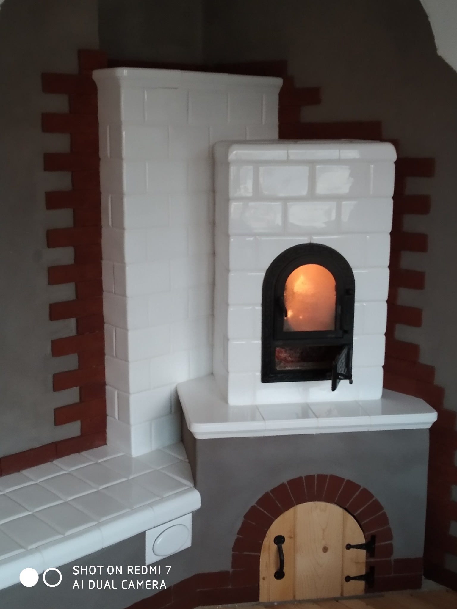Biały piec kaflowy z paleniskiem i drewnianymi drzwiczkami, obudowany cegłą, narożny, z widocznym płomieniem wewnątrz paleniska.