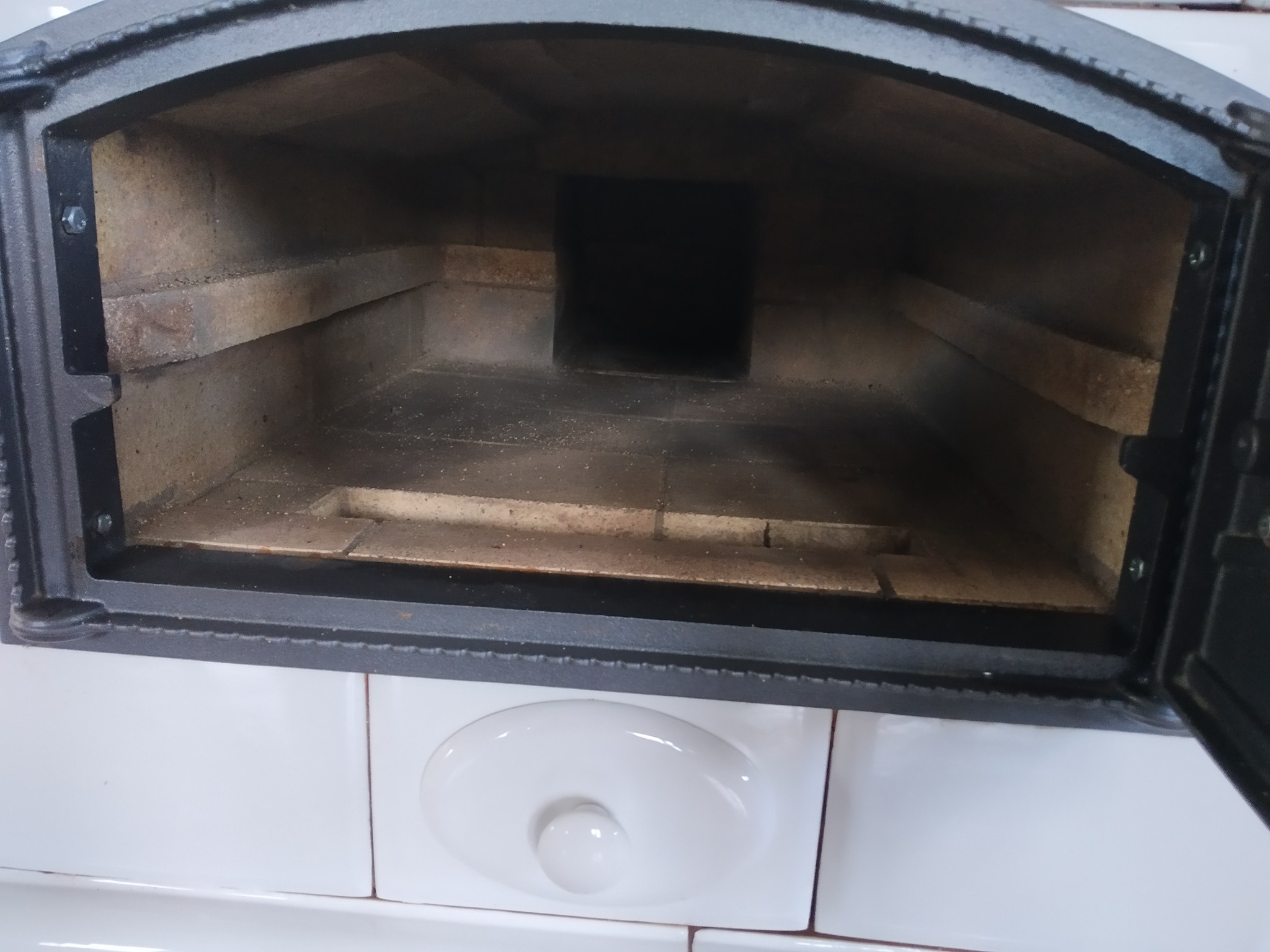 Otwarty piec kaflowy z czarnymi drzwiczkami i białymi kaflami poniżej, wnętrze wyłożone cegłą szamotową, widoczny komin. Detal pieca.