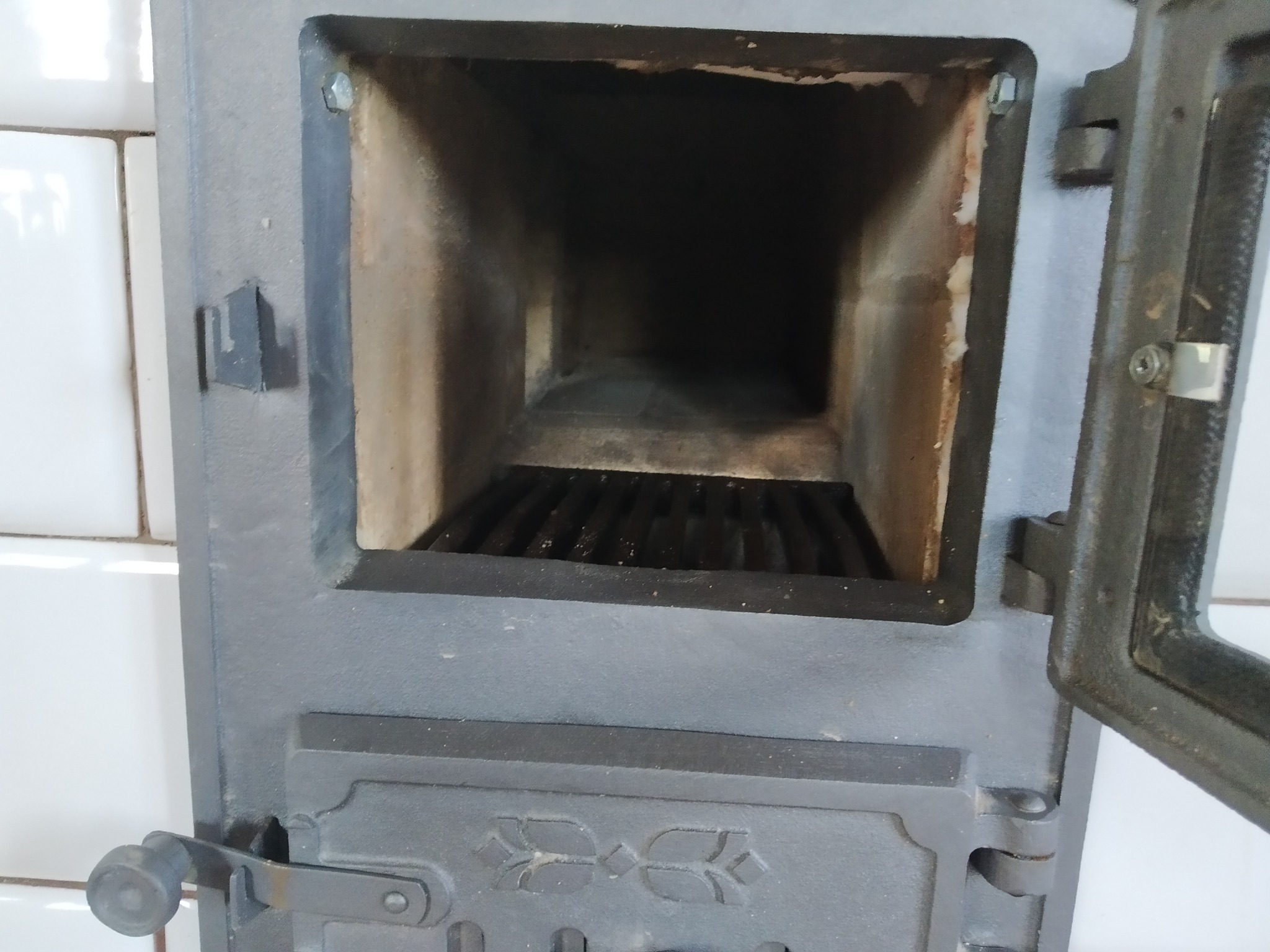 Otwarty, żeliwny piec kaflowy z rusztem wewnątrz, obok białe kafle. Widoczny mechanizm zamykający drzwiczki.