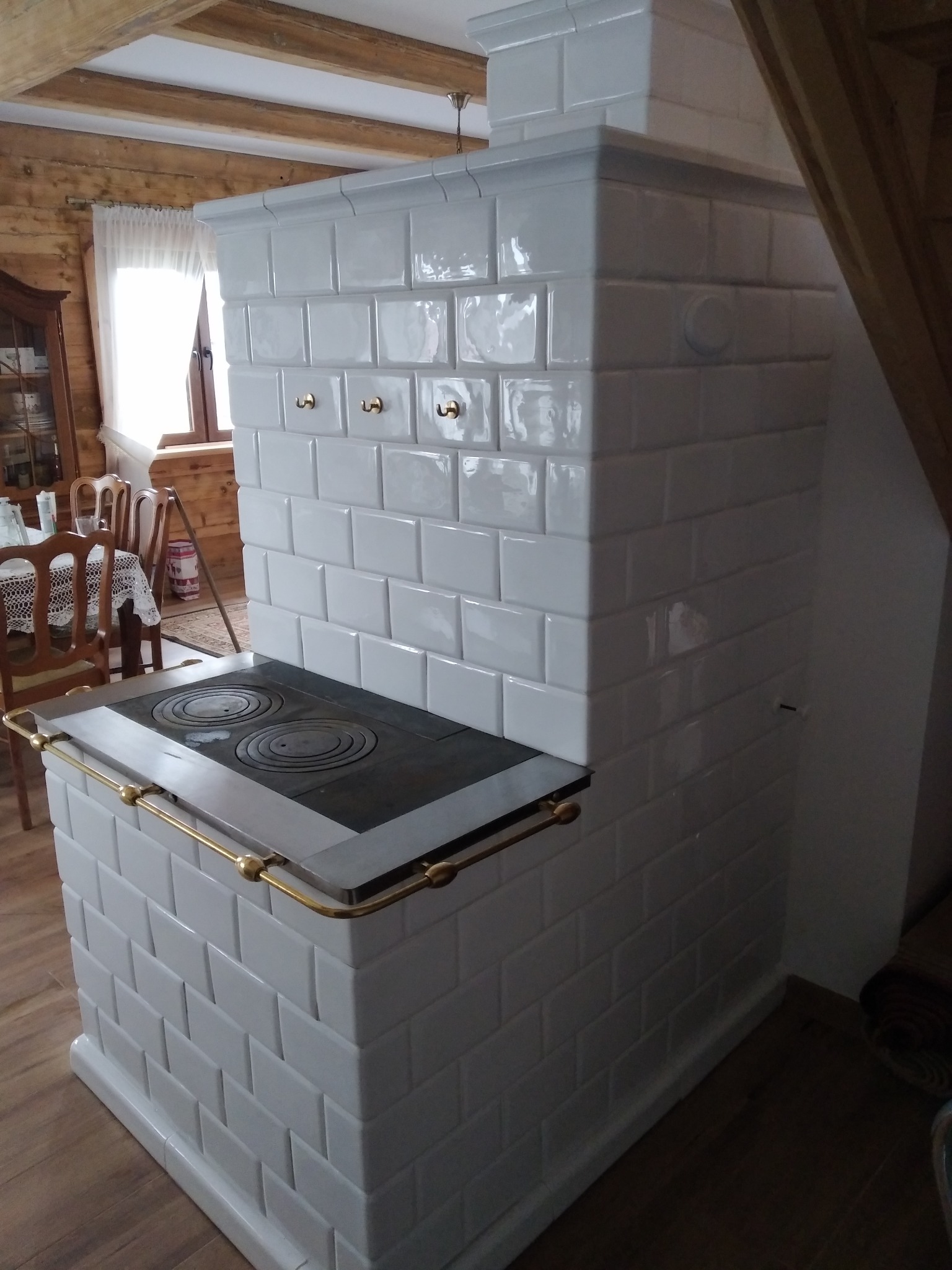 Biały piec kaflowy z płytą grzewczą i złotymi uchwytami, w rustykalnym wnętrzu z drewnianymi belkami stropowymi. Widoczne elementy wyposażenia kuchni.