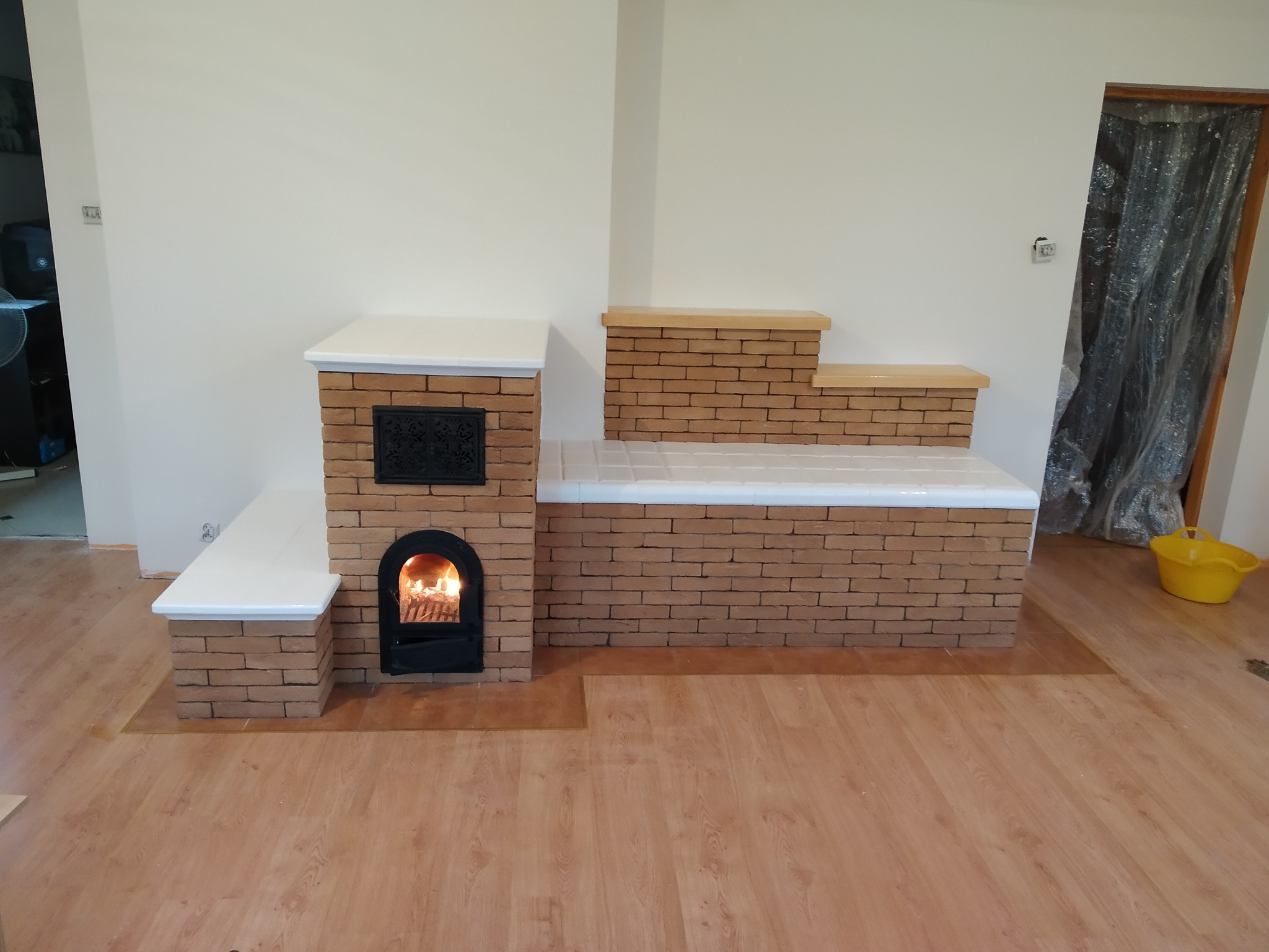 Oryginalny piec kaflowy z paleniskiem, obudowany cegłą klinkierową z białymi elementami wykończeniowymi. Widoczne płomienie w palenisku i drewniana podłoga.