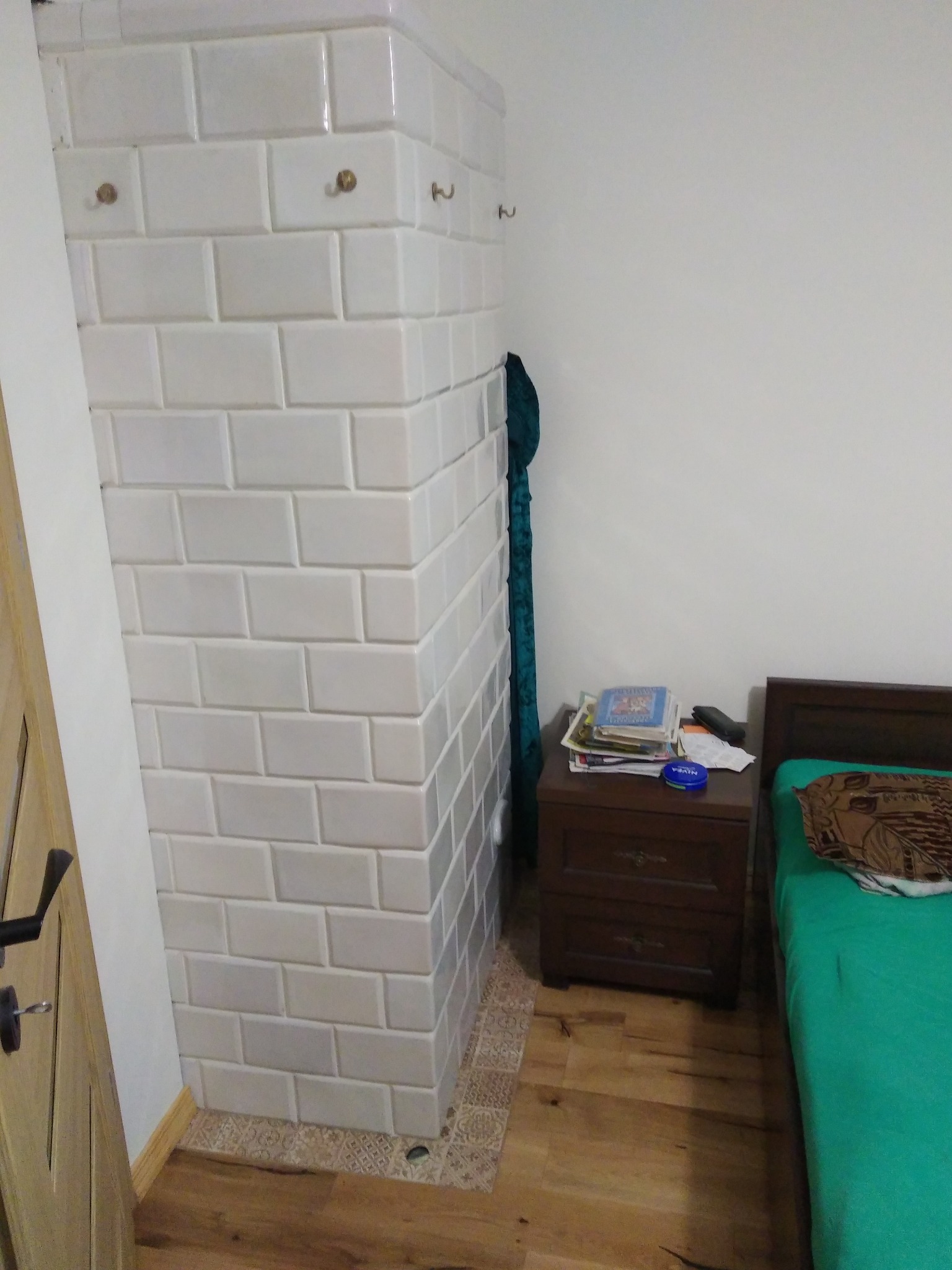 Biały piec kaflowy z haczykami w rogu pokoju, widok z boku, z elementami wystroju wnętrza: łóżko, szafka nocna, drzwi. Wykończenie podłogi z dekoracyjną bordiurą.