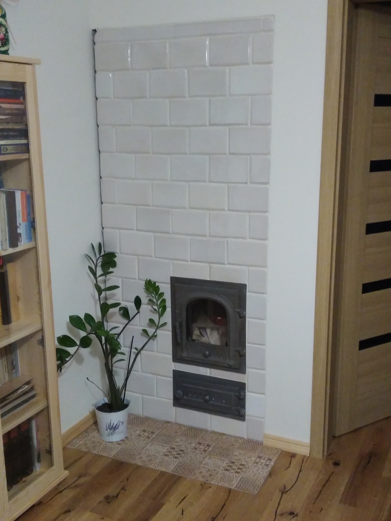 Biały piec kaflowy z żeliwnymi drzwiczkami, obok drewniana szafka z książkami i roślina doniczkowa, parkiet na podłodze, widok na drzwi.